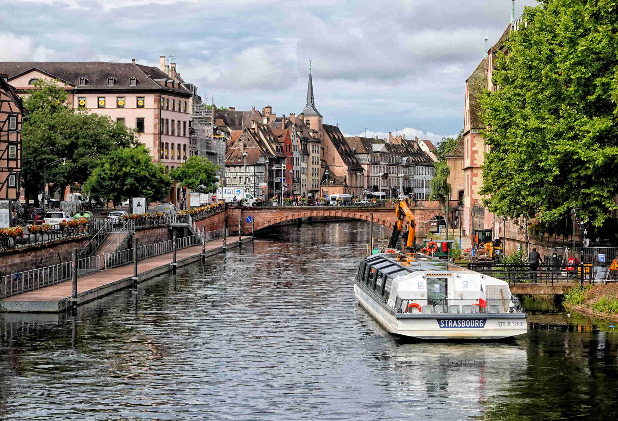 River running through a city, showing a bridge in the center, a passenger boat docked on the right shore, a passenger walkway on the left shore, and multi-story buildings along both sides