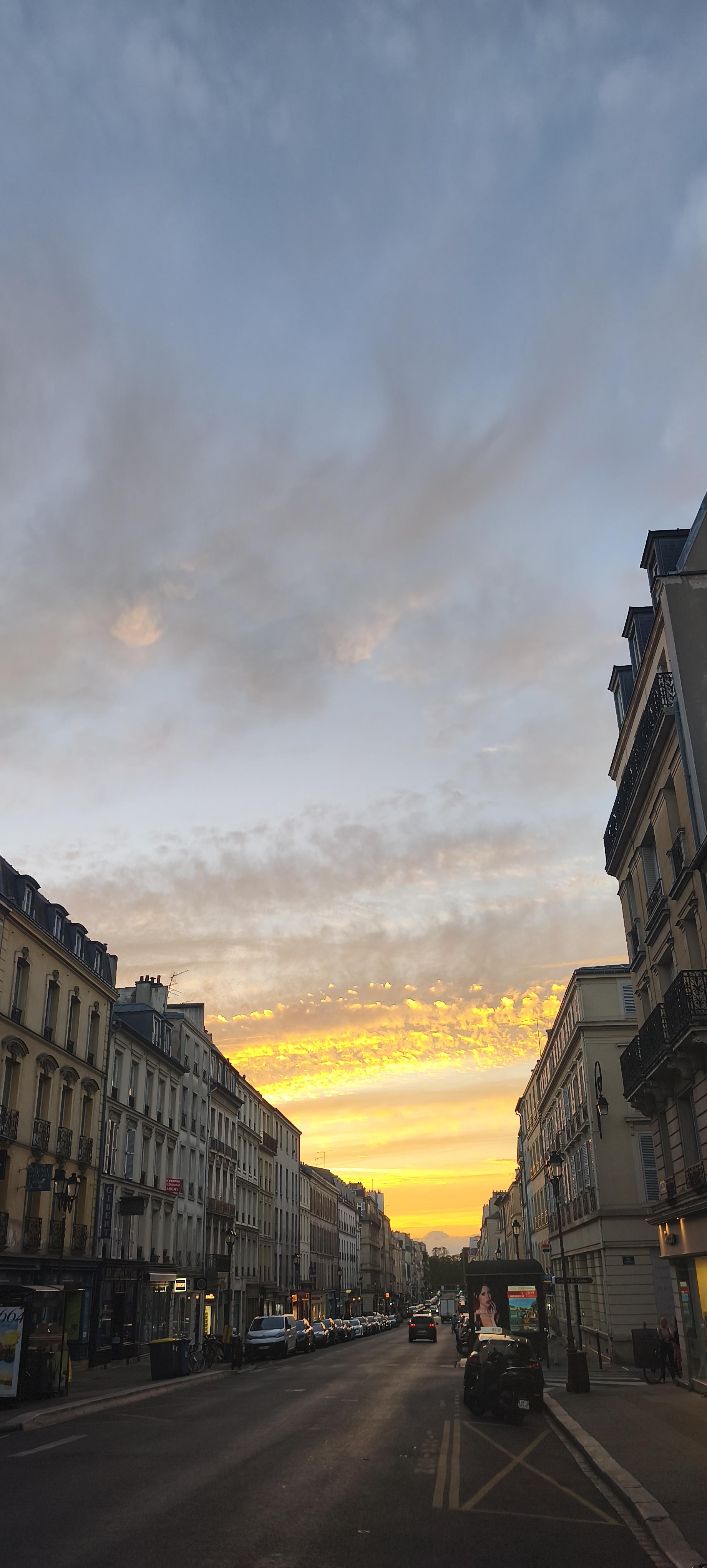 Rue versaillaise bordée d’immeubles, sous un immense ciel bleu-gris. À l’horizon, le soleil couchant embrase les nuages de jaune et d’or, illuminant le bout de la rue.