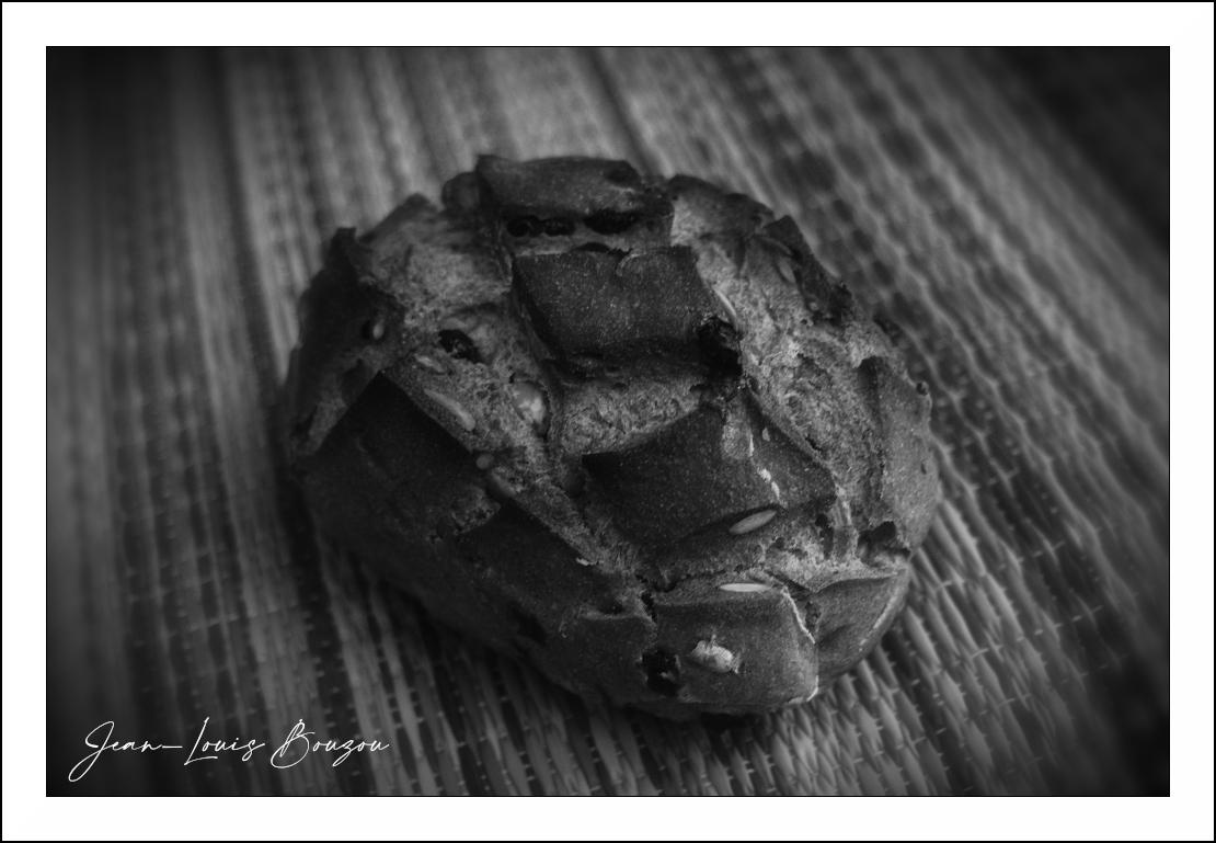 The image is a black and white, close-up, high-angle shot of a single, rustic loaf of bread. The bread is positioned slightly off-center to the right, dominating the foreground. It has a dark, textured crust with several scored lines, creating a crisscross pattern on its surface. Visible within the crust are inclusions that appear to be grains or seeds, as well as darker flecks that could be dried fruit or nuts. The texture of the bread suggests it is crusty on the outside and has a somewhat dense crumb.

The bread remains on a woven mat or placemat. The mat's pattern consists of parallel, textured lines that run diagonally from the upper left to the lower right of the frame. The weave of the mat creates a repeating textured background that contrasts with the organic form of the bread.

The lighting in the image creates strong contrasts and highlights the texture of both the bread and the mat. The overall tone is monochromatic, with shades of gray ranging from deep, dark shadows to bri