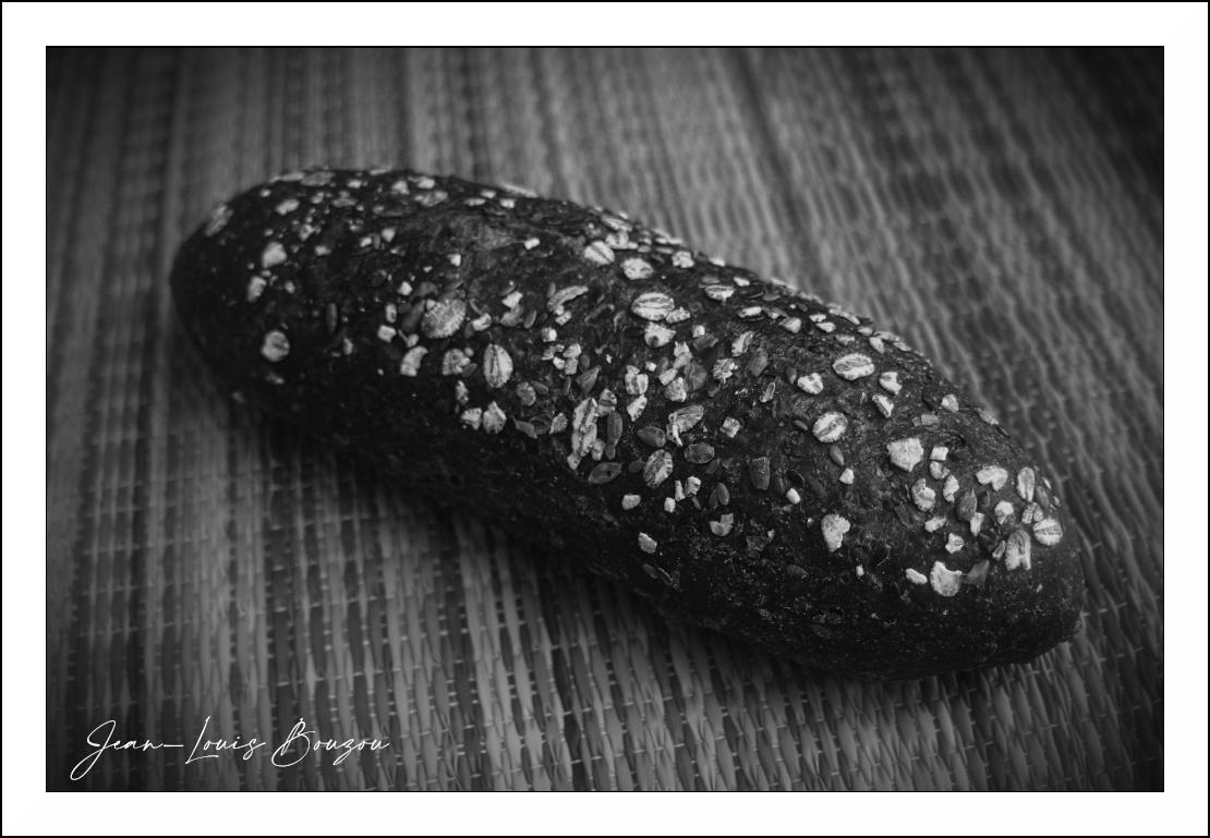 The image is a black and white, close-up, high-angle shot of a dark, rustic loaf of bread lying diagonally across a woven mat. The bread is elongated and oval-shaped, with a slightly uneven surface. It is generously topped with various seeds and grains, including visible oats, sunflower seeds, and possibly flax seeds. These toppings create a textured and speckled appearance against the dark crust of the bread. The bread occupies the majority of the frame, extending from the bottom left towards the upper right.

The mat beneath the bread has a distinct woven pattern, appearing as a series of raised lines and indentations that create a subtle texture and shadow play. The lines of the mat run roughly parallel to the long axis of the bread. The background is softly out of focus, drawing attention to the detail of the bread.

The color palette is monochromatic, consisting of shades of gray, black, and white, which accentuates the textures and forms within the image. The lighting appears to 