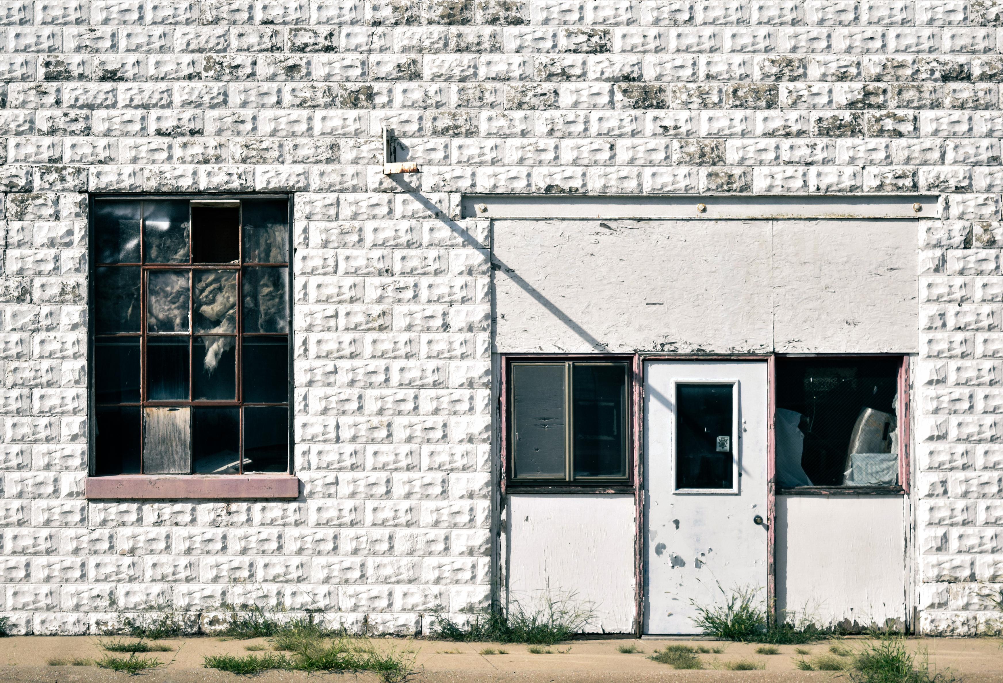 Weathered white brick building with a barred window and worn double doors, cracked glass, and peeling paint, evoking a stark, abandoned mood.