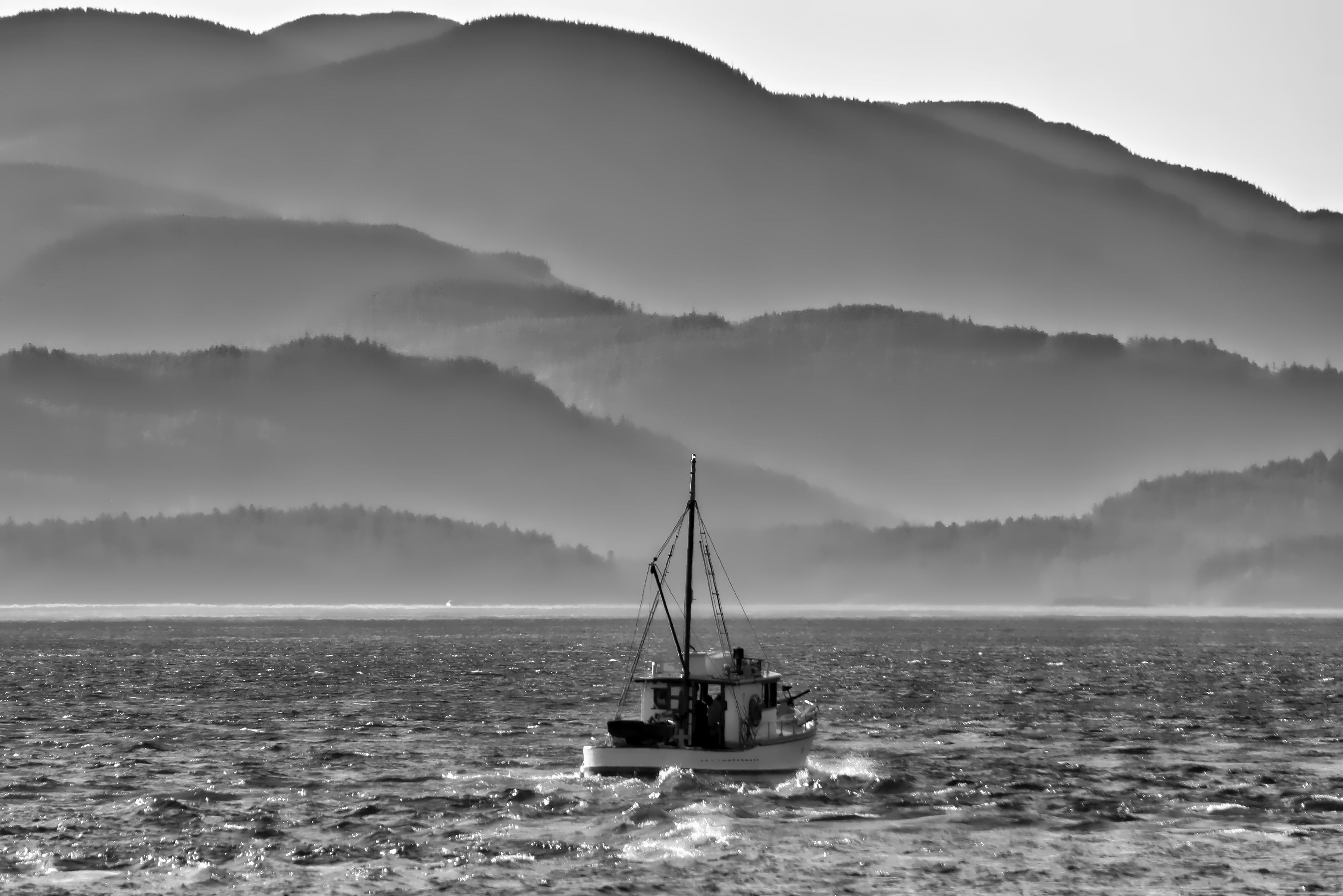Schwarzweiss-Foto eines kleinen Fischerbootes vor der hügeligen Küste. Die Höhenzüge der Küste liegen noch etwas im Nebel.