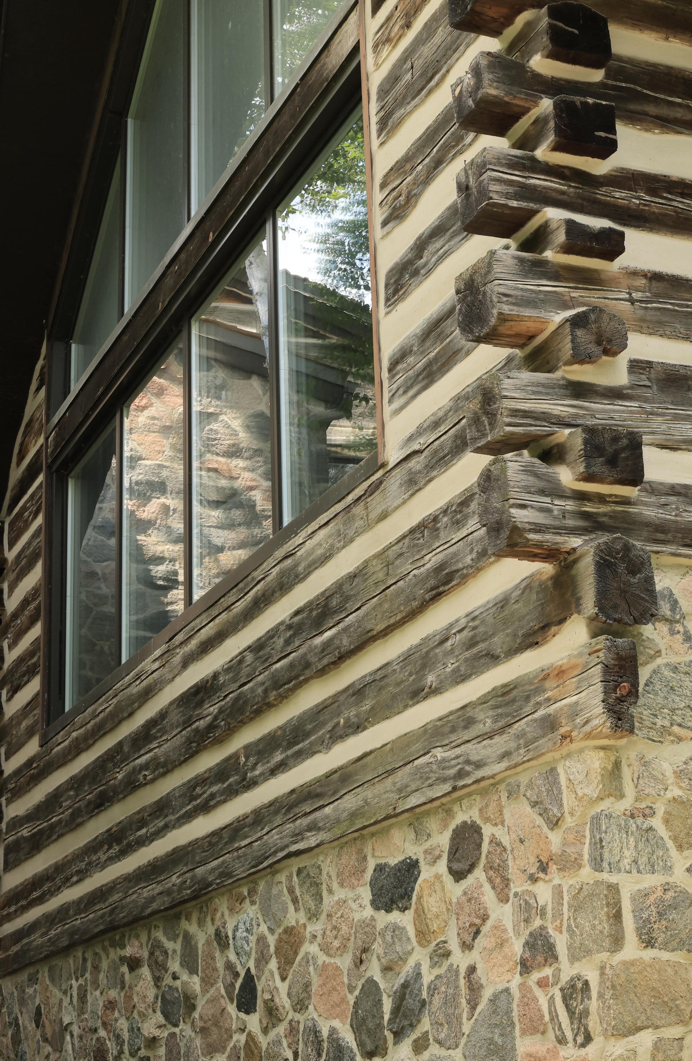 This is a portrait format photo taken in the summer of one wall of a "log built" building. The lower section of the wall is constructed of stonework, with squared off logs making up the upper portion. A larger window in the wall shows the reflection of a stone chimney which is on an adjacent wall. The corner of the wall shows how the squared  off logs notch into each other.  Lots of texture in this photo.
