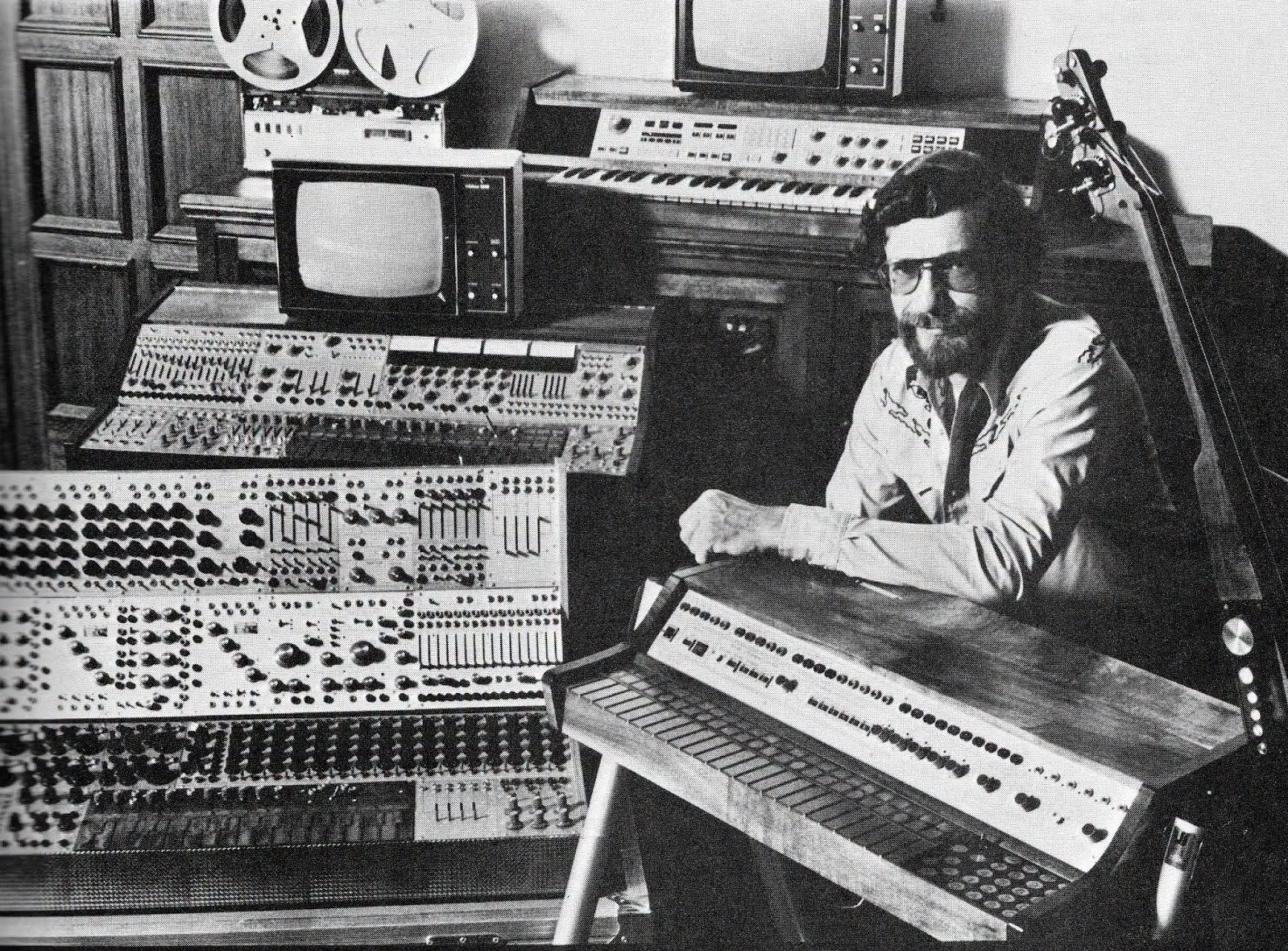 American electronic music pioneer Don Buchla posing alongside his early modular synthesizers.