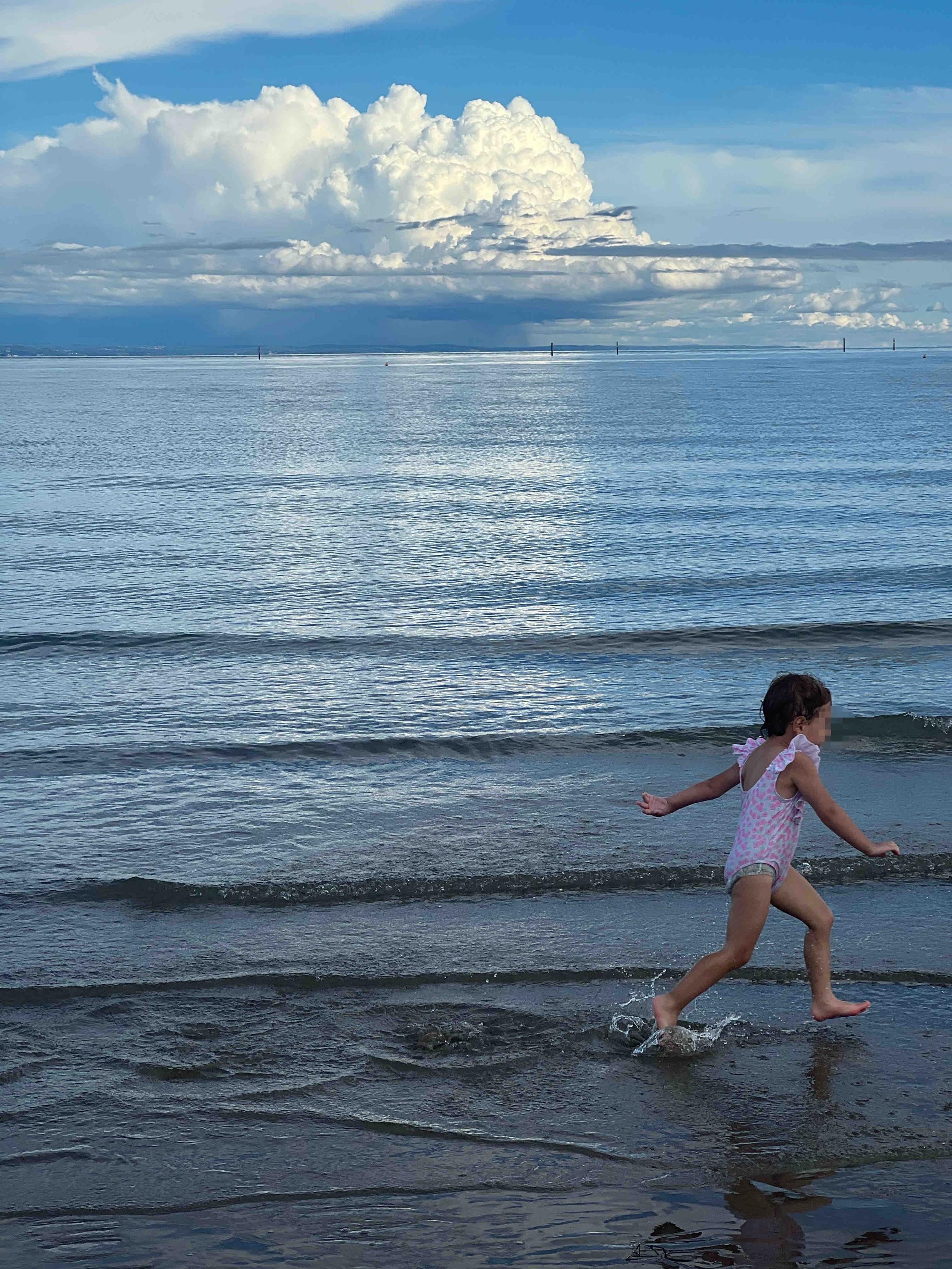 Hochformat Aufnahme. Ruhiges Meer. Am Horizont eine Cumulus Wolke und Regen. Im Vordergrund hüpft ein Mädchen durch das flache Wasser am Strand

Portrait format photo. Calm sea. At the horizon a cumulus cloud and rain. In the foreground a little girl runs through the shallow water at the beach