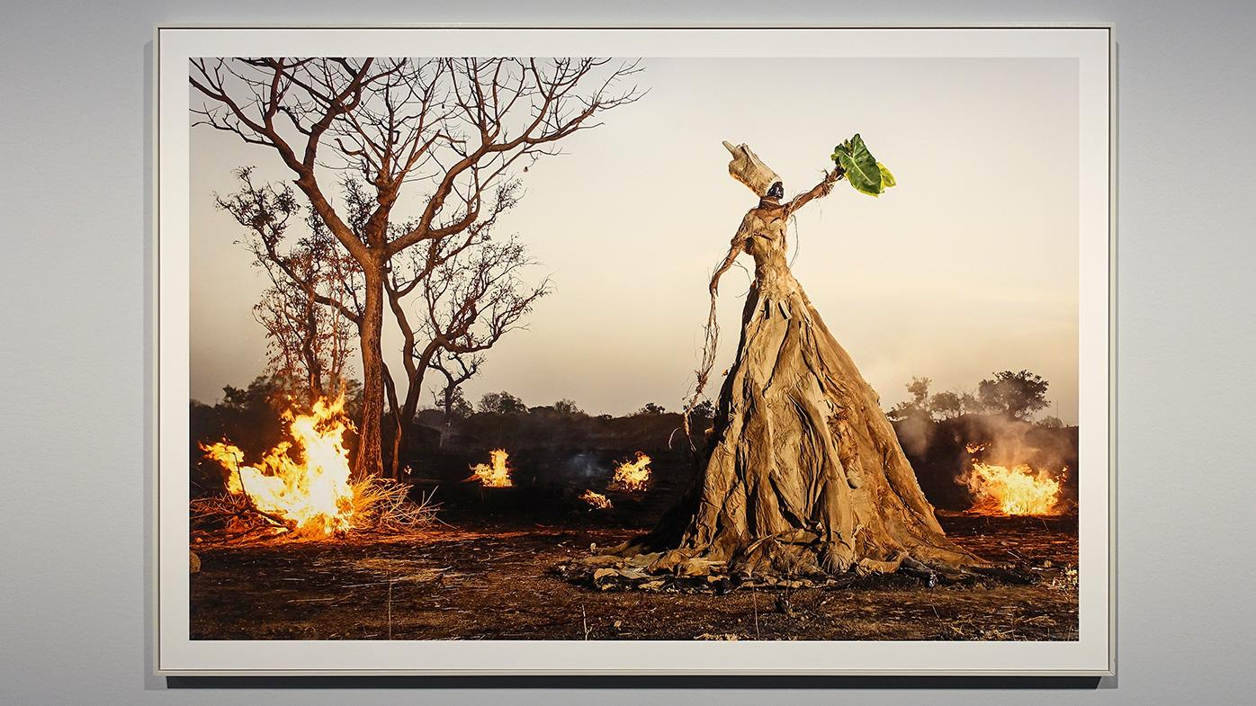Kleurenfoto van een landschap met bomen en struiken tijdens de avondschemering. het en der vuren. Een donker persoon is gekleed in een jurk van bruin afval en is vele meters hoog. Op haar hoofd heeft ze ook een koker van hetzelfde afval. In haar hand houdt zij twee groene palmbladeren vast en richt deze naar de hemel. Alsof de goden worden gewaarschuwd. Het geheel ziet er dreigend uit.