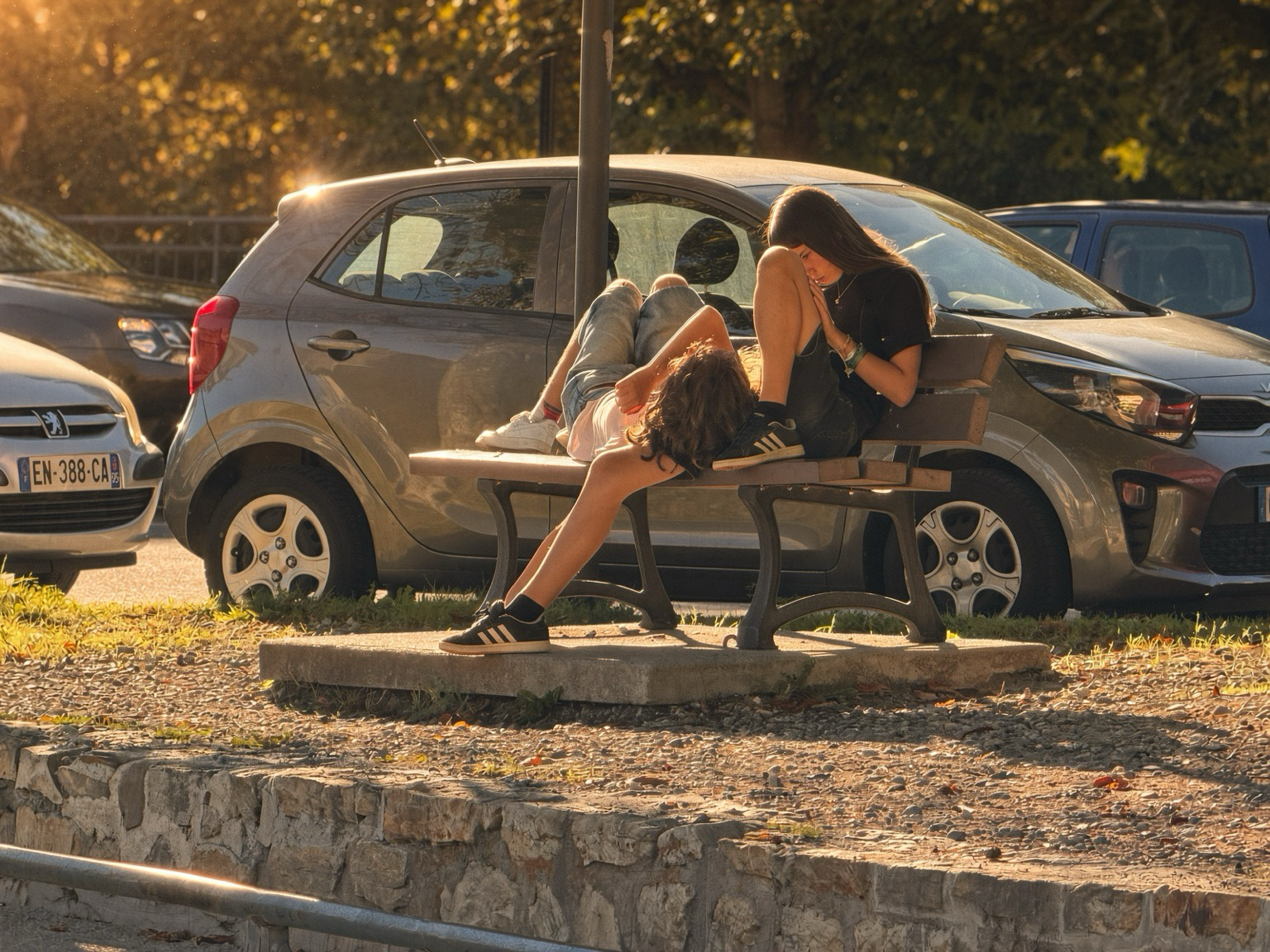 Two young people sit on a wooden park bench in a parking lot during golden-hour sunlight. One person lies across the bench in denim shorts and white sneakers, head near the other person’s lap. The second person sits upright with knees drawn up, wearing a black T-shirt, dark shorts, black Adidas sneakers, and a green wristband, looking down at a phone. Behind them is a grey Kia hatchback, other parked cars, trees, and warm low sunlight. A low stone wall and gravel occupy the foreground.
