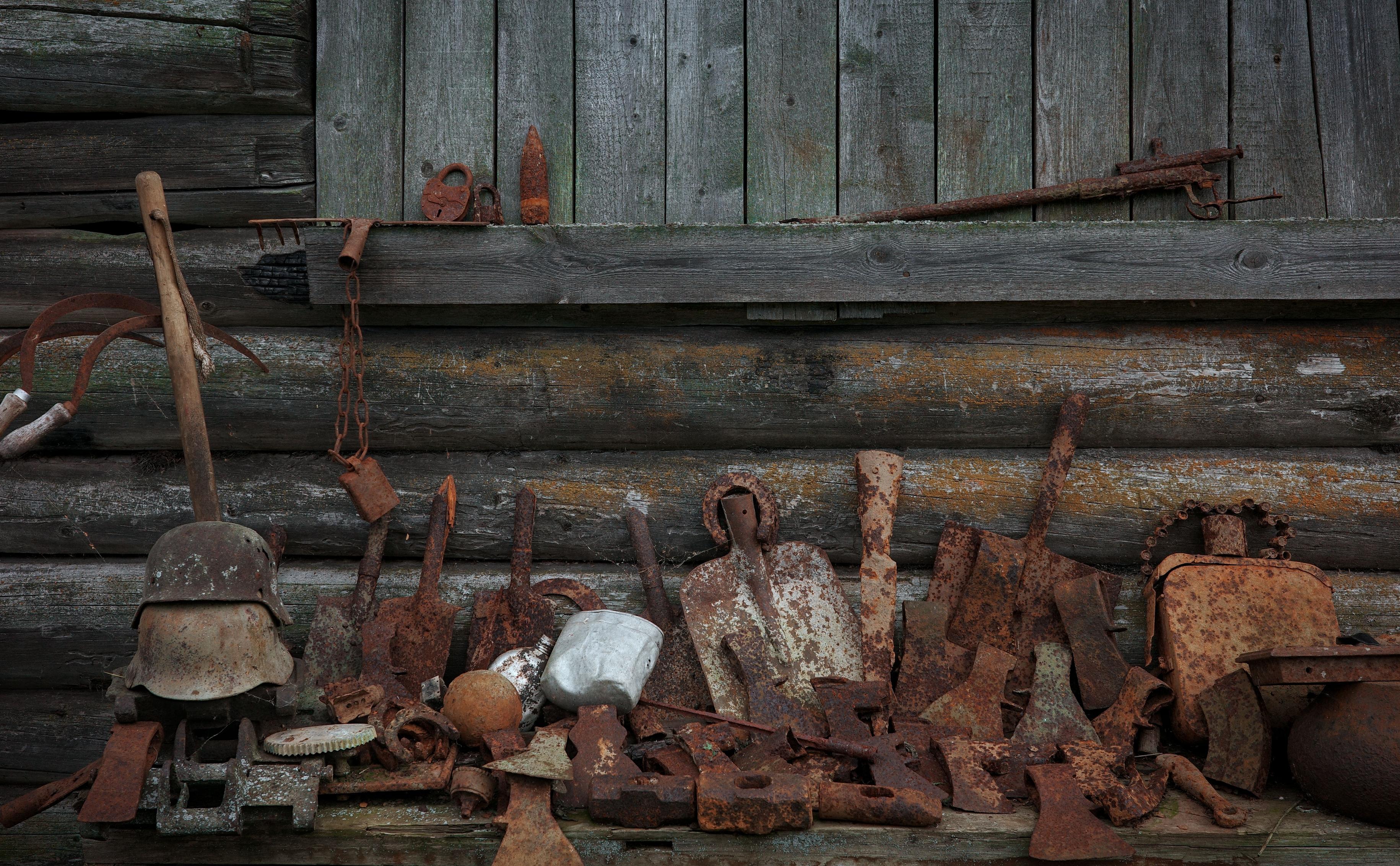 This color photograph captures the wall of an old wooden log house, turned into an impromptu exhibition of old, rusted objects. The upper part of the composition consists of even, gray-green logs with a horizontal shelf, on which lie a rusty padlock, a cartridge case, and an old rifle. The lower part is a dense pile of metal artifacts: a German helmet on a stick, rusty bayonets of shovels, crowbars, axes, and fragments of machinery and household items. On the left, a white metal can and a rusty sphere stand out. This entire heap is covered in deep, warm rust, contrasting with the cold wood. The image conveys a feeling of abandonment, memory, and the harsh beauty of bygone eras.
