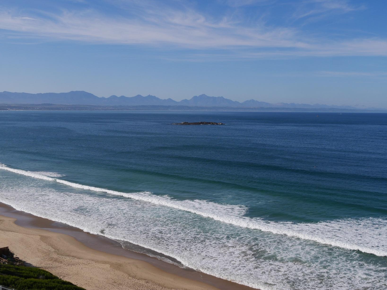 Der helle Sandstrand von Mosselbay trifft auf den grünblau schimmernden südlichen Ozean. Nahezu die Schnittstelle zwischen Atlantik und Pazifik. Im Hintergrund sind Berge erkennbar. Südafrika 2022