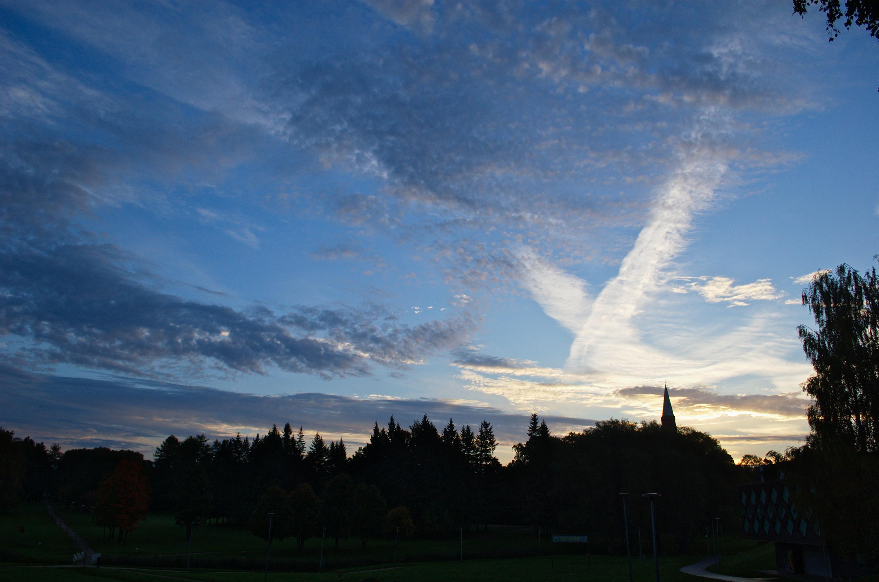 Photo of a blue sky partially covered in scattered dark clouds. There’s a brighter band on the horizon, lit by the recently risen sun. The clouds on the right side of the scene are mostly white, dominated by two wide diffuse contrails, crossing each other in an X shape.