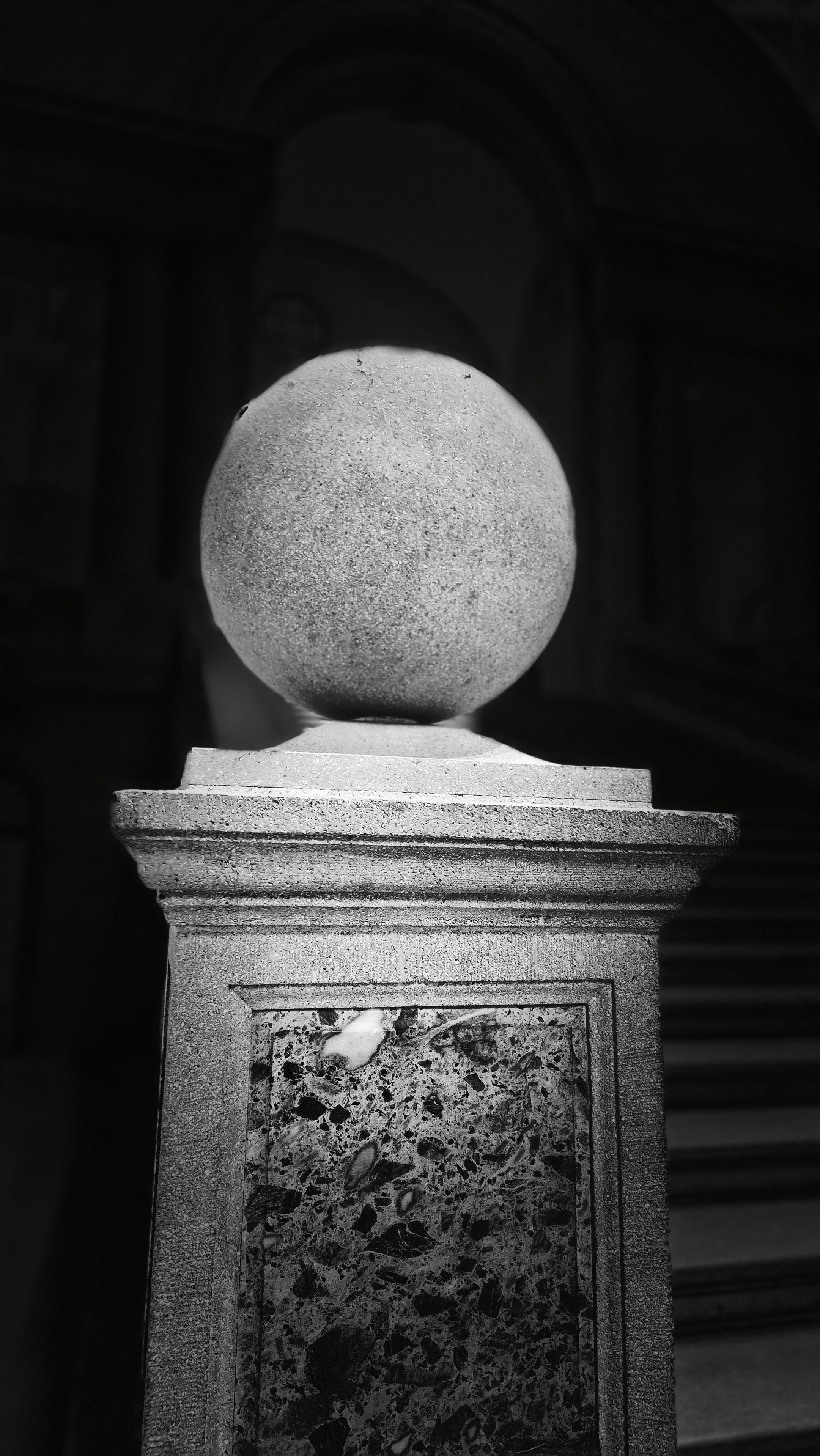 Nahaufnahme einer Kugel auf einem Sockel in schwarzweiß. Im Hintergrund sind ein Treppenaufgang und Säulen schwach zu erkennen. /
A close-up of a sphere on a plinth, in black and white. A flight of stairs and columns are faintly visible in the background.