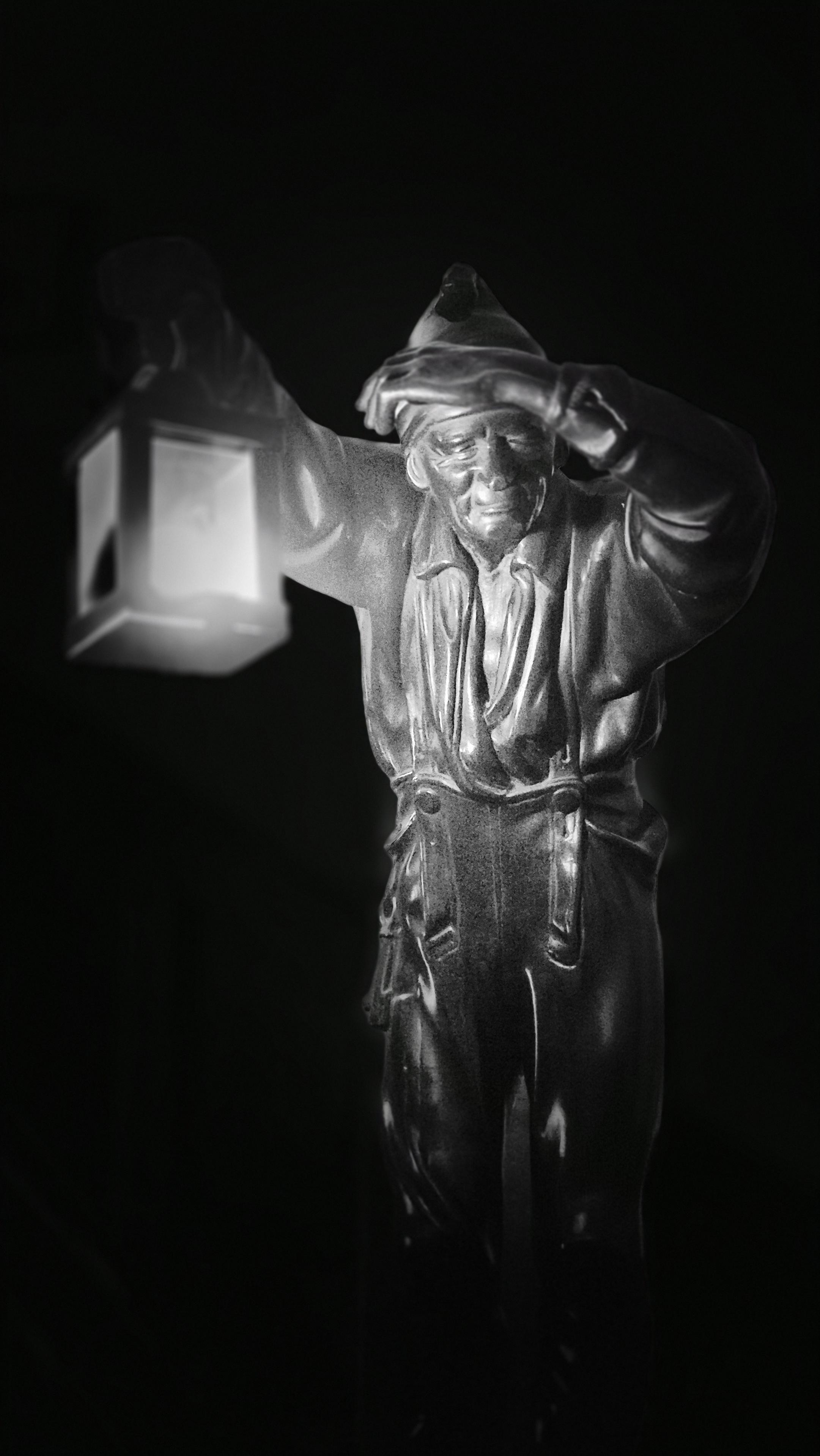 Nahaufnahme einer Holzfigur, die sich die linke Hand vor die Stirn hält und eine leuchtendene Laterne in der rechten trägt, vor schwarzem Hintergrund. Das Foto ist in schwarzweiß. /
A close-up of a wooden figure holding its left hand to its forehead and carrying a glowing lantern in its right hand, against a black background. The photograph is in black and white.