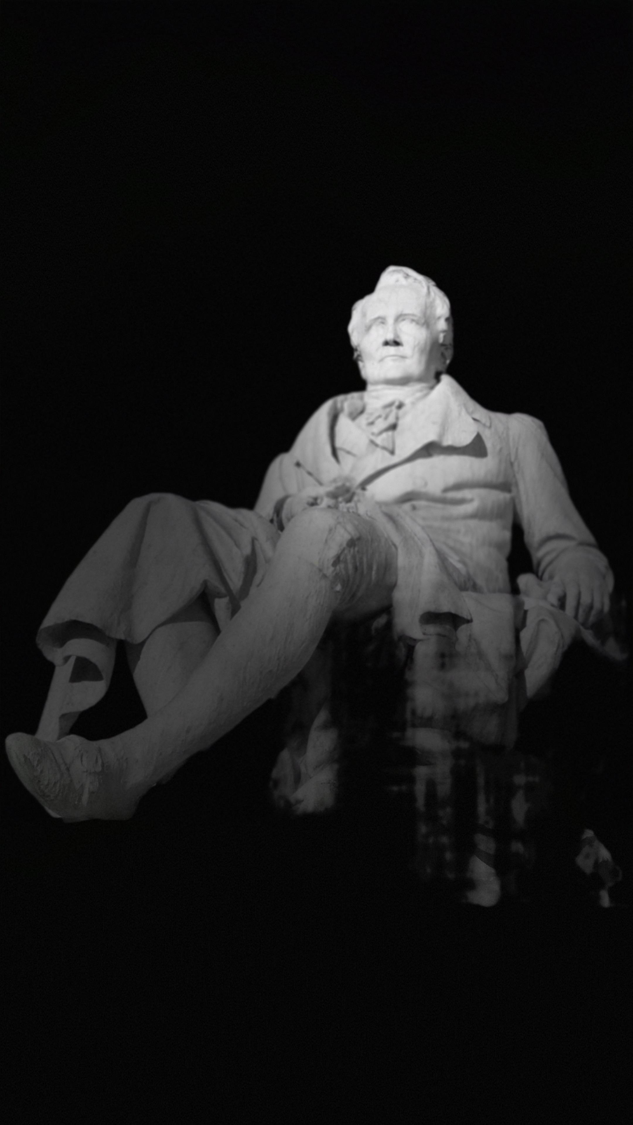 Das Schwarzweißfoto zeigt die schwebende Büste eines Alexander von Humboldt in einer Nahaufnahme vor schwarzem Hintergrund. /
The black-and-white photograph shows a close-up of Alexander von Humboldt’s floating bust against a black background.