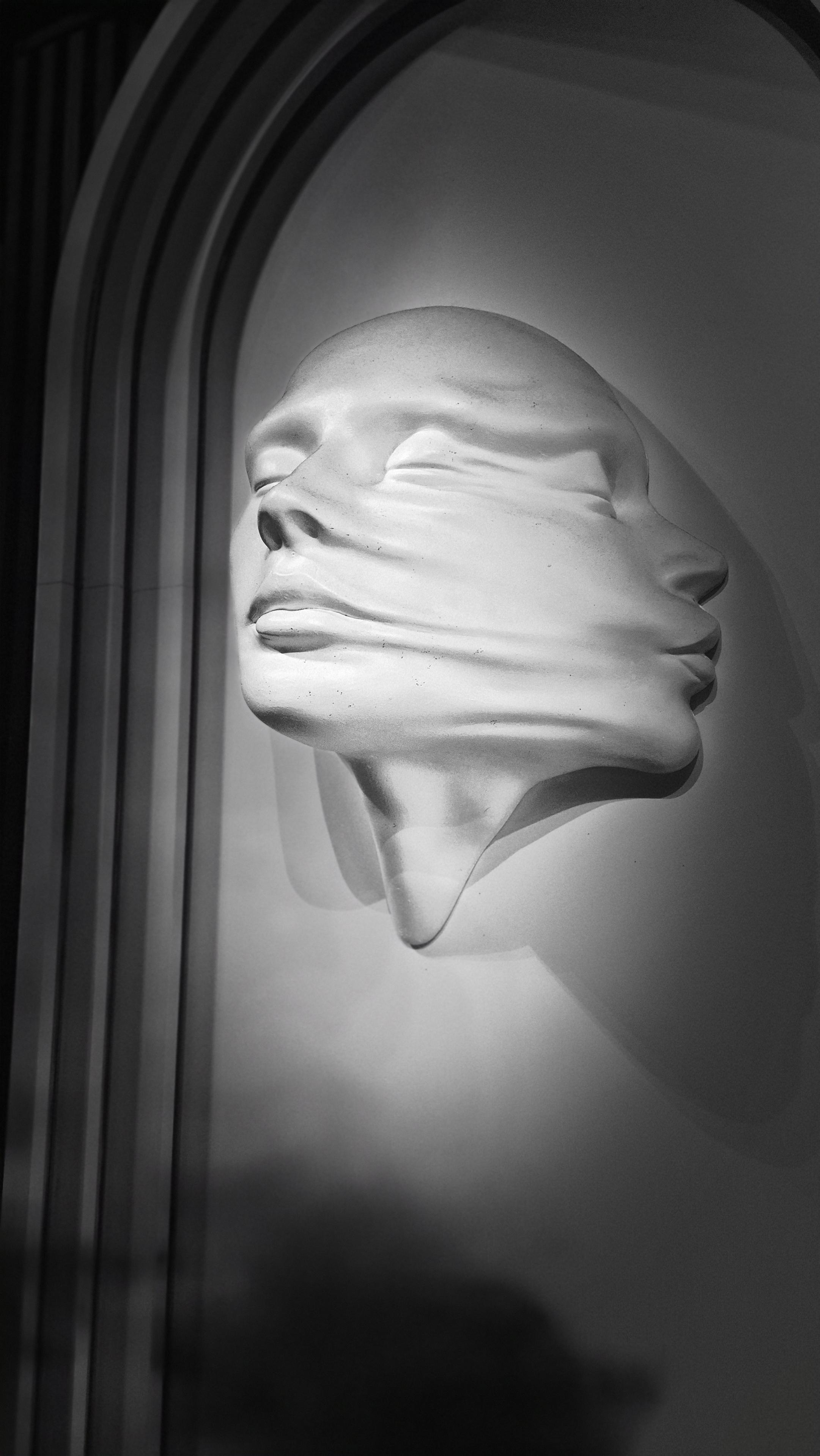 Nahaufnahme zweier auseinanderdriftender, verzerrter Gipsköpfe, die an einer Wand hängen und Schatten werfen, in schwarzweiß. /
A close-up of two distorted plaster heads, drifting apart, hanging on a wall and casting shadows, in black and white.