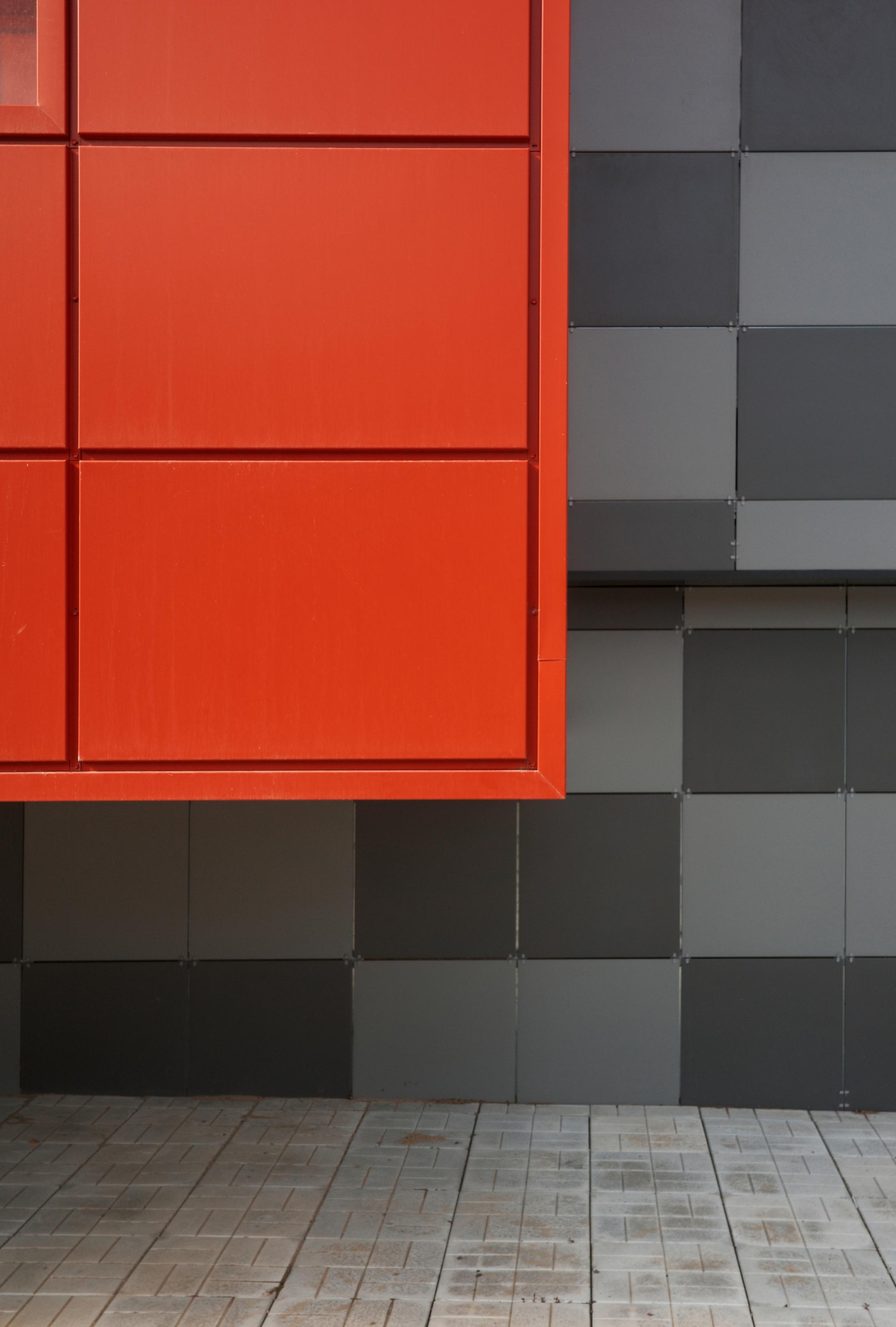 This color photograph captures a fragment of a modern facade turned into an abstract geometric composition. In the left part of the frame, a large red block with clear horizontal divisions dominates, resembling an insert panel or a billboard. It sharply contrasts with the right part of the wall, which is clad in square panels of various shades of gray, creating the effect of a chessboard or pixel mosaic. Below is paved tile with texture and traces of time. The image is built on pure colors, flat shapes, and a strict rhythm of lines, turning an ordinary wall into a minimalist, almost abstract canvas.