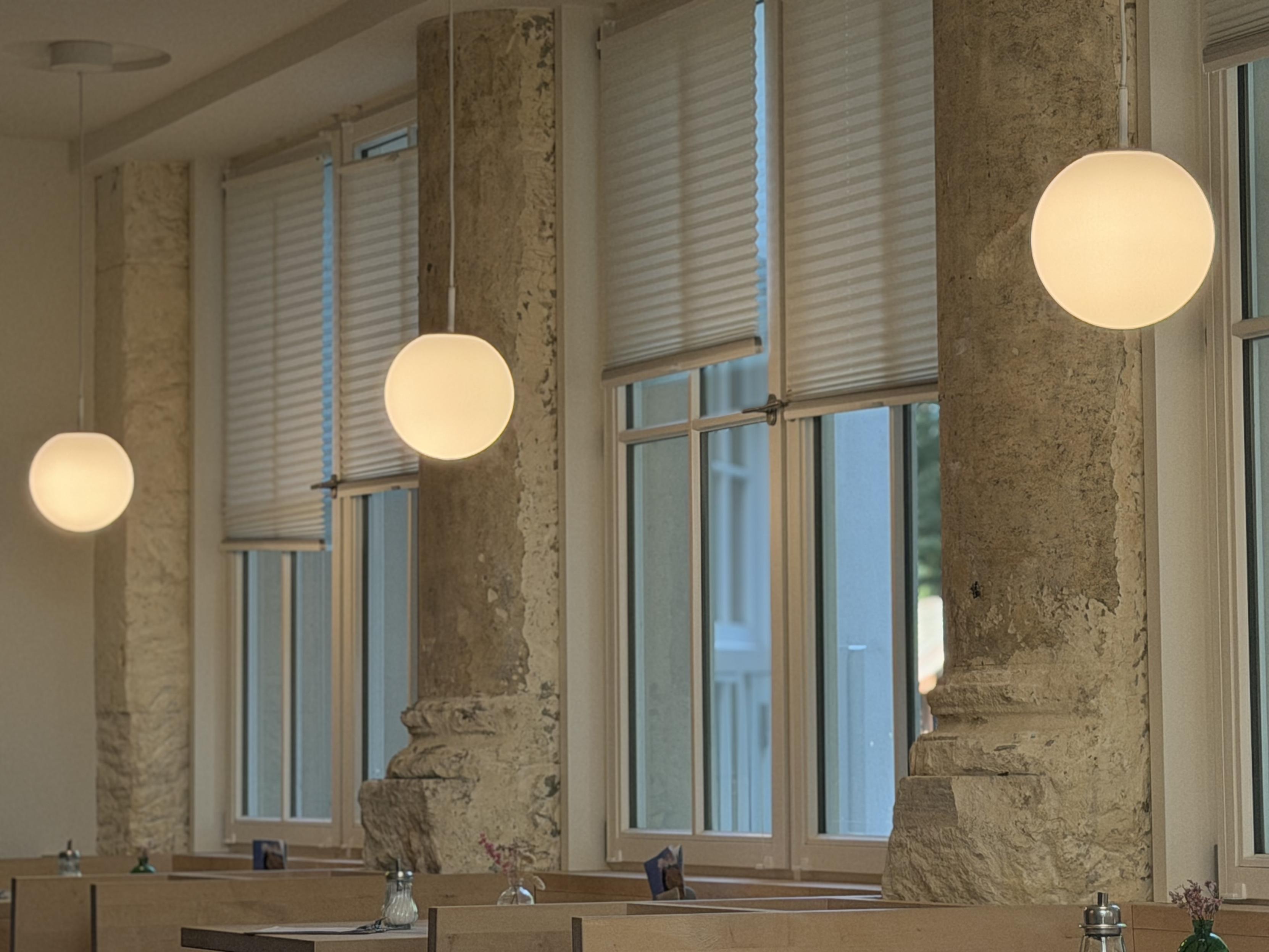 Interior view of a restaurant or café with tall mullioned windows, some with pleated blinds partially lowered. Three spherical, glowing white pendant lamps hang from the ceiling at varying heights, arranged in a row. Between the windows stand massive, roughly plastered sandstone pillars with molded bases. In the lower part of the frame, wooden tables are set with salt and pepper shakers and small flower vases.