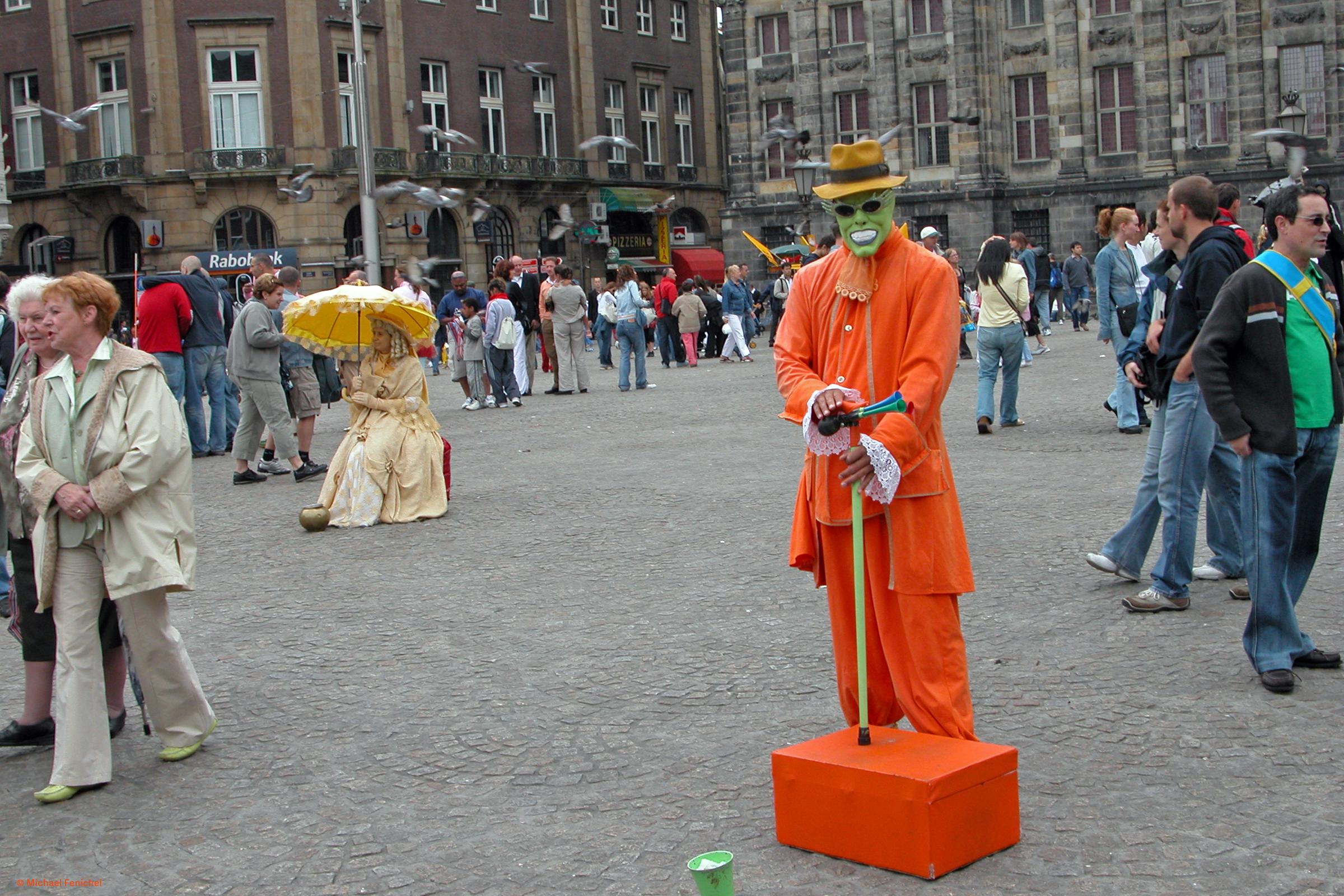 Photo by Fenichel: 
Dam Square in Amsterdam, in mid-day. With an orange character from "The Mask" in center, on the cobblstone, passersby all around, and to the left a woman in yellow sits on a chair with a yellow parasol. Behind are brick/stone buildings.
