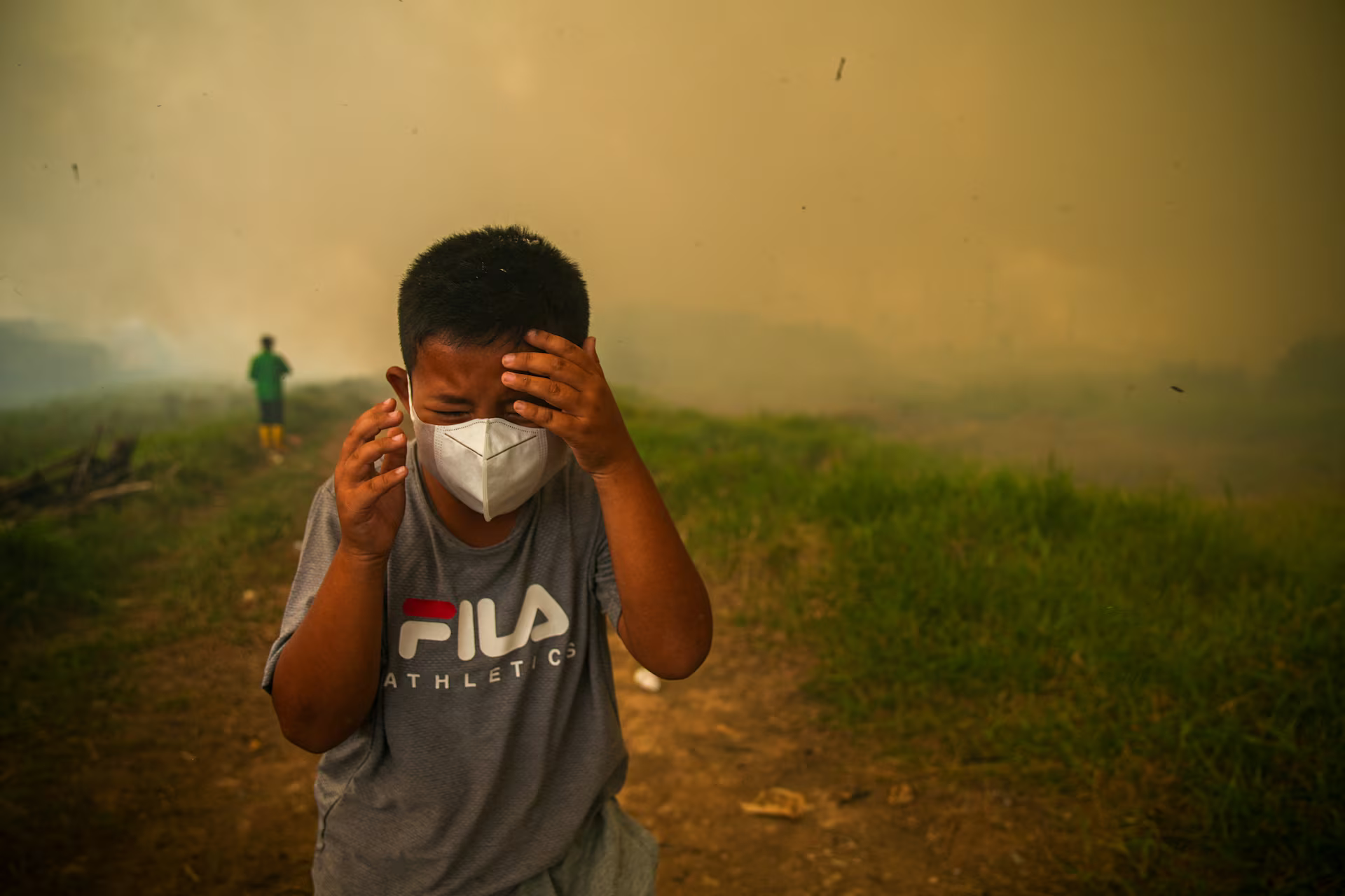 A boy with a face mask grimaces as smoke rises from wildfires.