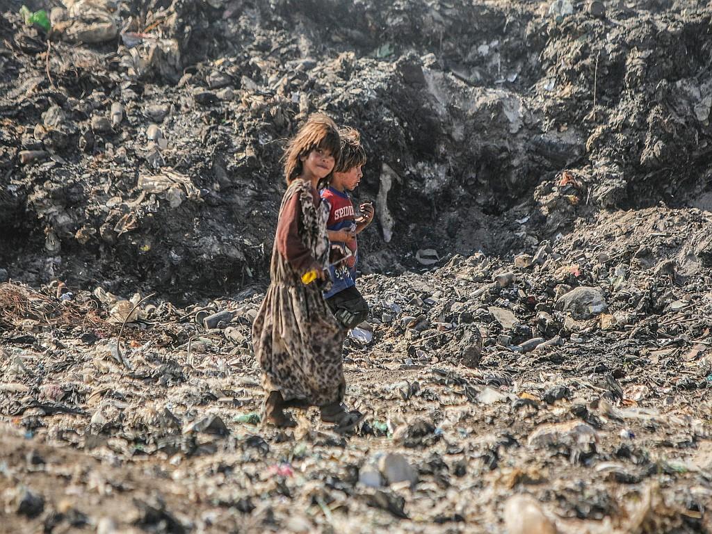 A girl & a boy pass a mountain of rubbish at a dump.
