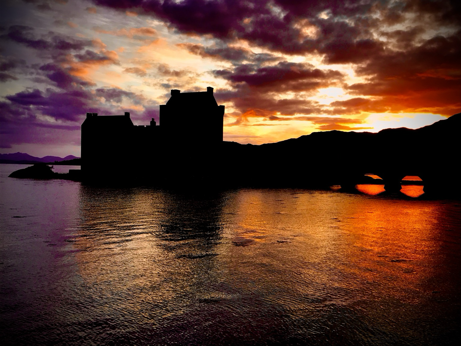 The sun setting behind a silhouetted Eilean Donan castle in Lochalsh in the Scottish Highlands. The water ripples gently as it flows under the access bridge. The sun paints the sky in mixtures of red, orange, yellow, purple and blue.