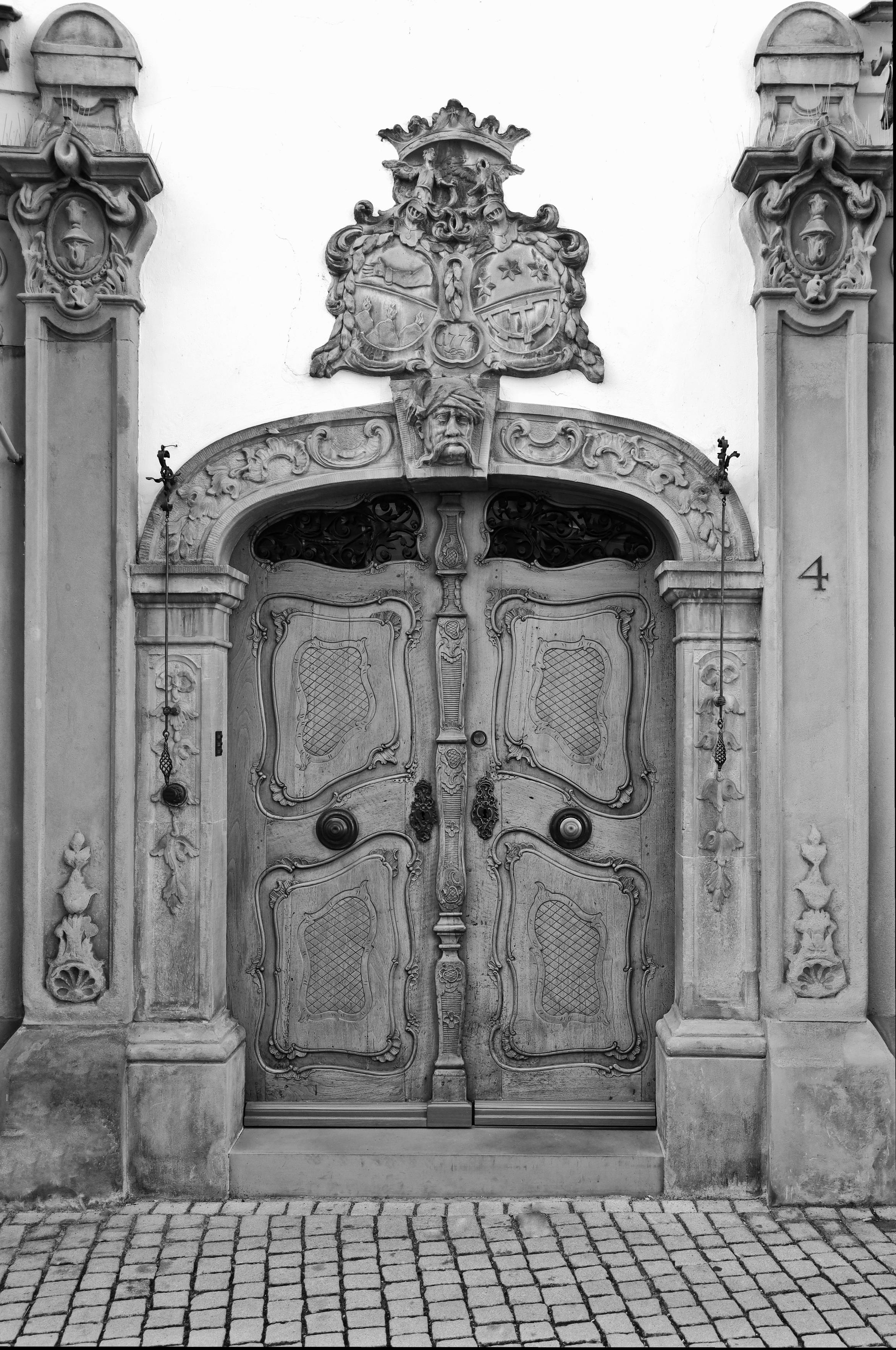 Schwarzweiss-Foto einer hölzernen, historischen Haustür in Konstanz am Bodensee, die von Elementen aus Stein umfasst ist. Unten vor der Tür ist kleines Kopfsteinpflaster zu sehen.