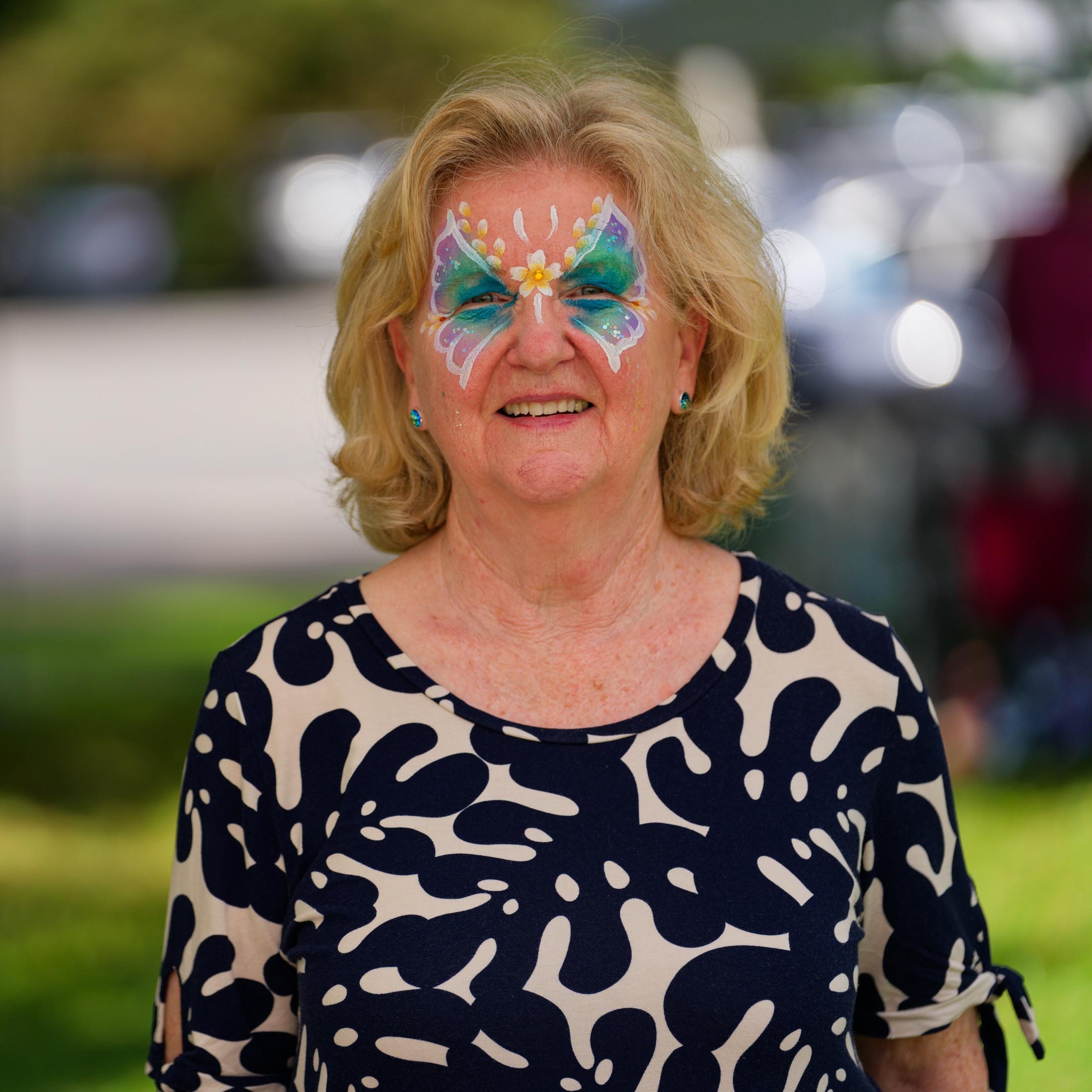 Older woman with blue and white butterfly painted around her eyes, medium blonde hair and a black and beige top