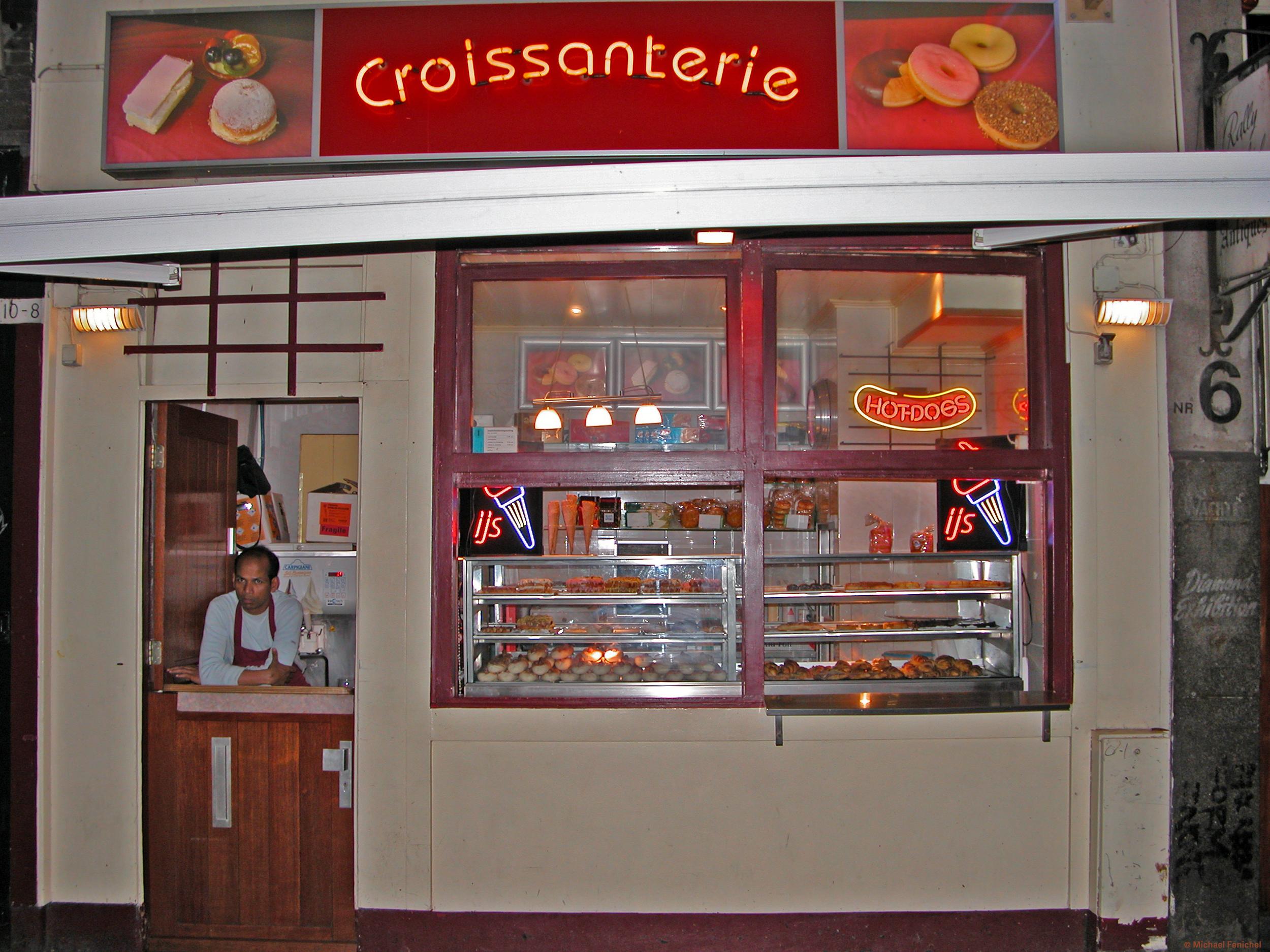 Photo by Fenichel:  "Croissanterie #6"

An image of a small shoppe with a door and half-window on the left (through which orders are taken) and four large windows (2x2) to the right.

The shop is beige with red/brown wooden trim, and the top foot/meter or so is a strip of store name/advertising, with a red center pannel and neon yellow-orange lettering: "Croisanterie" . To the left and right of "Croissanterie" are a panel with illustrations of cookies, donuts, and pastries.

Back at the ground level, we see a man in apron leaning on the open window (1/2 door) on the left. In the windows to the right are rows of pastries, and some hot foods, including "Hot Dogs", for which there is neon sign inside a hot dog shaped 'bun", upper right window. Far right of the shop, a sliver of wall has a sign - NR 6 - and a lamp.