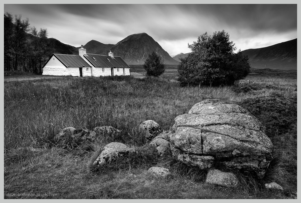 A photograph of Blackrock Cottage standing alone in the rugged landscape of Glencoe, Scotland, with the imposing slopes of Buachaille Etive Mòr rising dramatically behind it. The small cottage has whitewashed walls that stand out against the darker, wild surroundings, while the long exposure and black and white treatment create a calm, stark atmosphere across the scene.