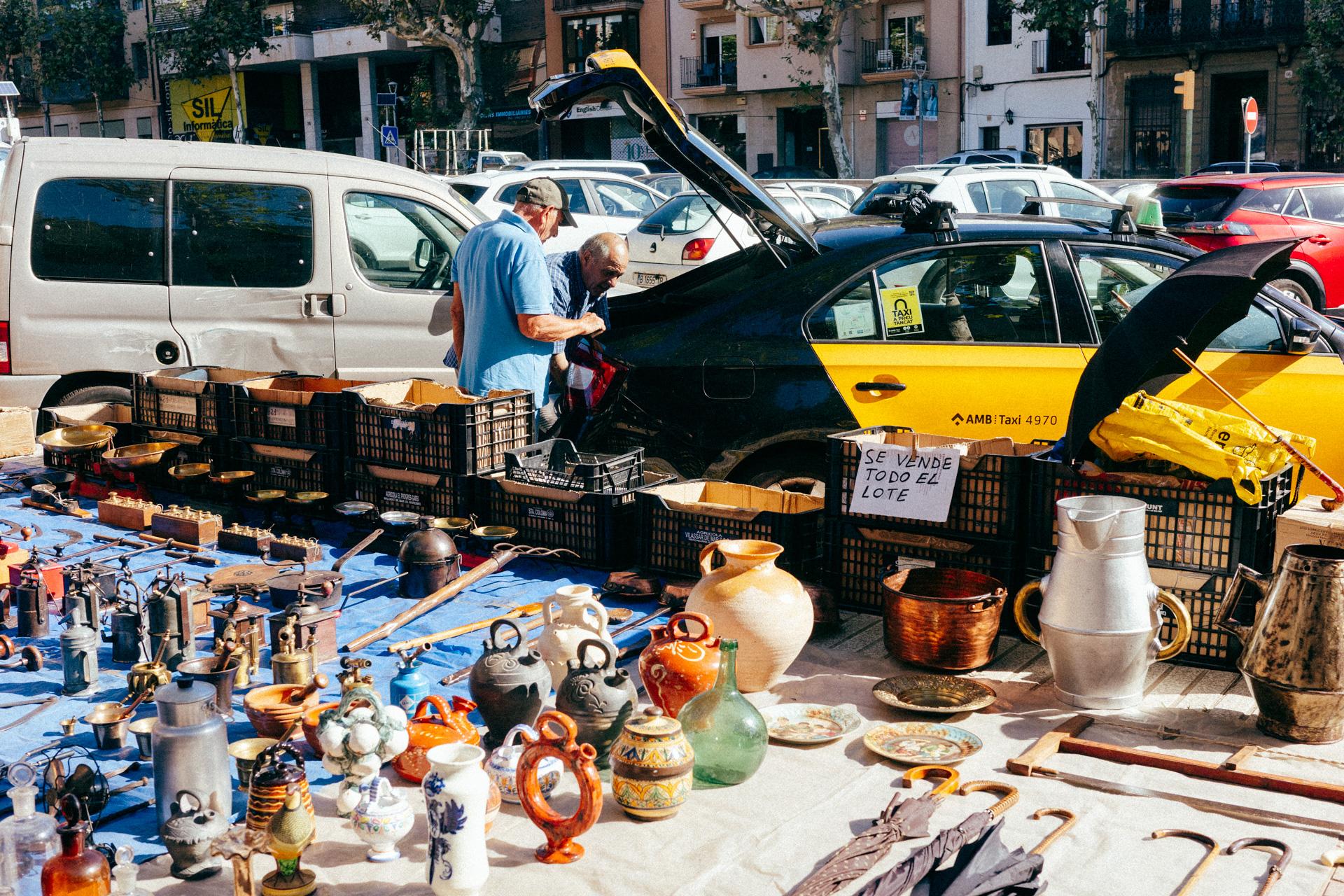 Fotografía de un puesto del mercadillo de Arenys de Mar lleno de herramientas y objetos curiosos. Al fondo hay coches aparcados y dos personas haciendo tratos en el capó de un taxi. También se puede leer un cartel escrito a mano que dice "se vende todo el lote".
A photograph of a stall at the Arenys de Mar market, filled with tools and curious objects. In the background, there are parked cars and two people making a deal over the hood of a taxi. A handwritten sign reading "entire lot for sale" is also visible.