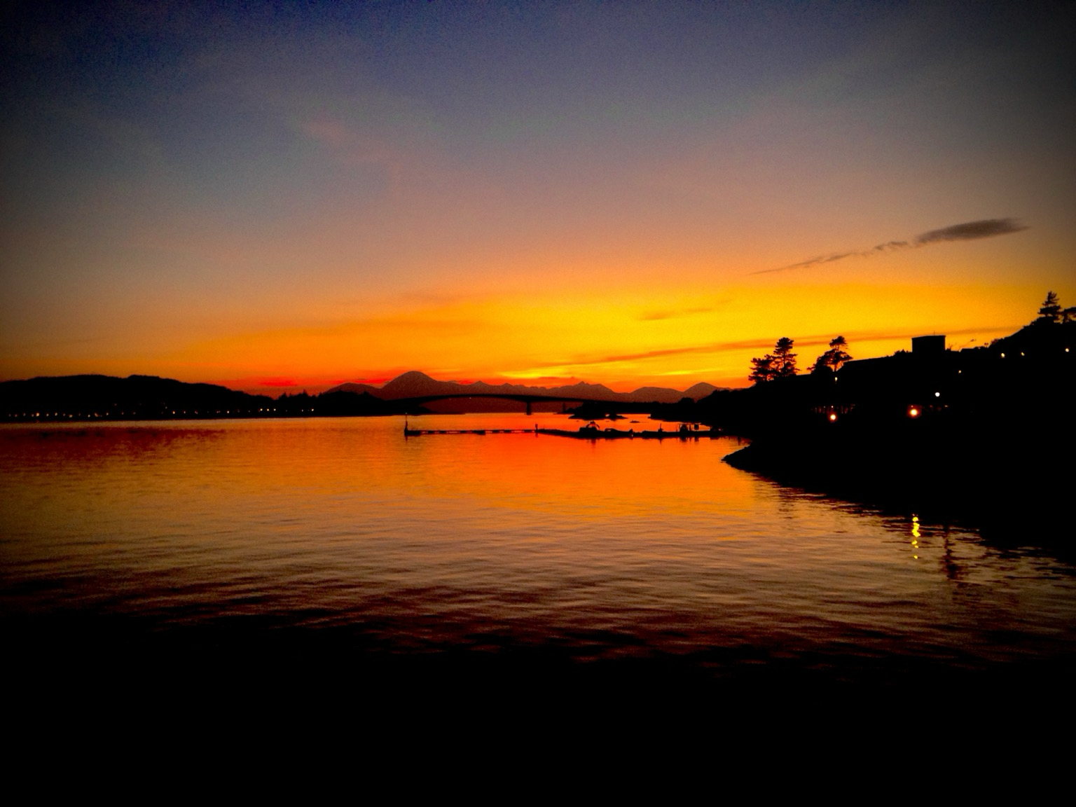 From the railway platform at Kyle of Lochalsh, the sun sets over the Isle of Skye and the Cuillin mountain range. The sky is a burnt orange melting into a muted blue, reflected in the dark but calm waters.
