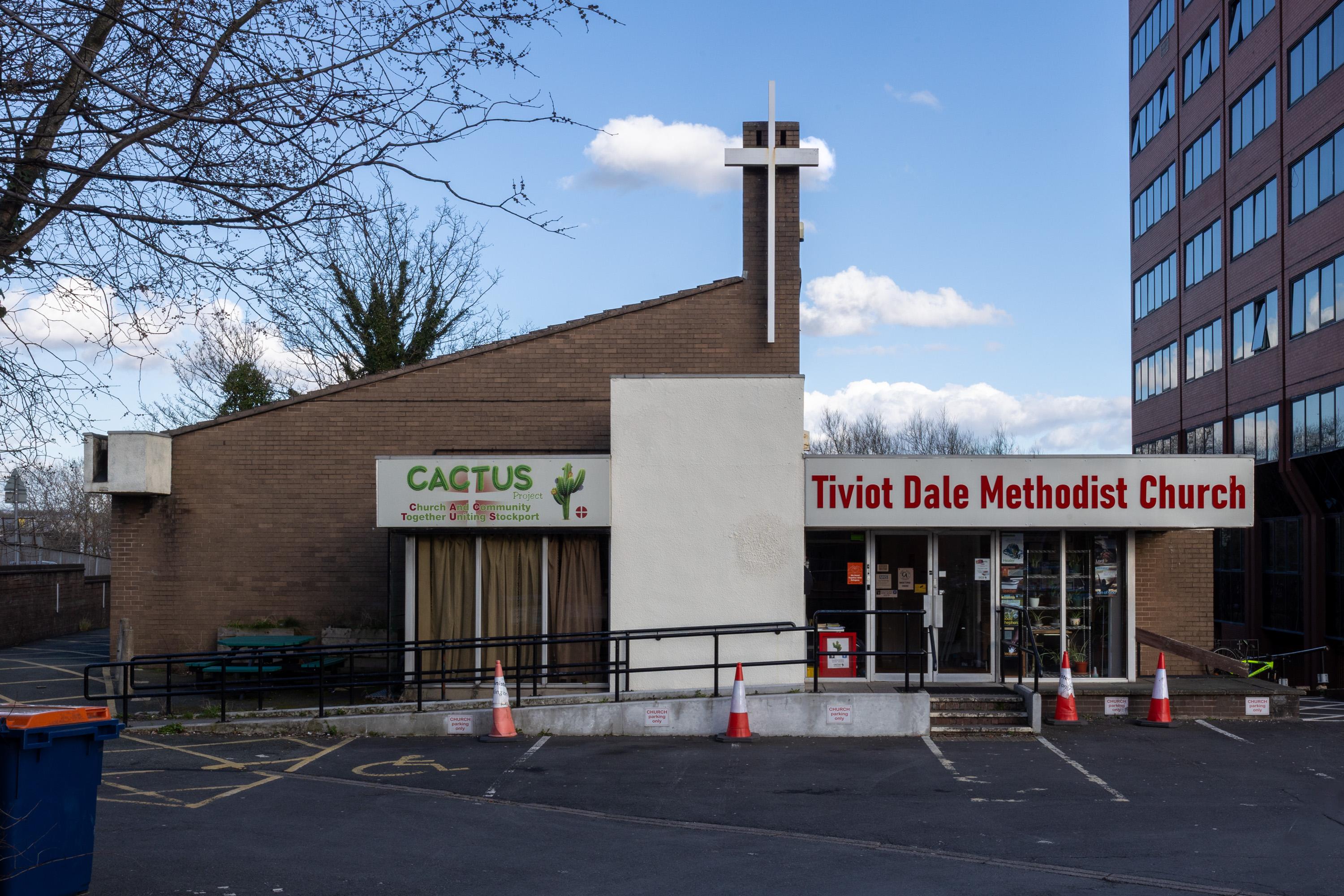 A brick building houses Cactus Café and Tiviot Dale Methodist Church. A cross tops a chimney. Orange cones line a ramp and car park. A tall office block stands to the right. Leafless tree branches frame the left side.