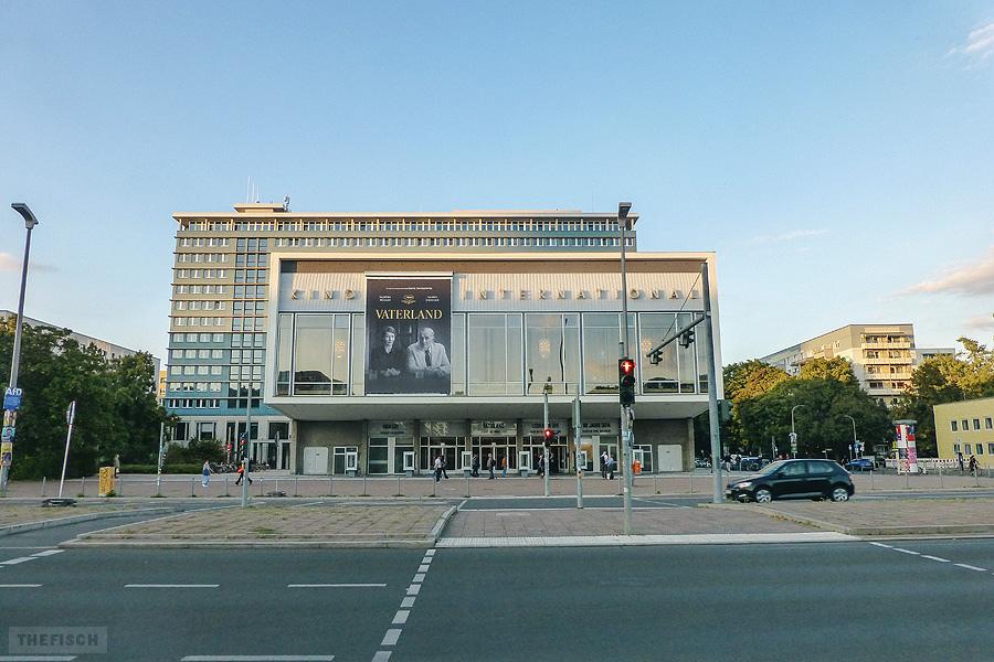 Das Kino International in Berlin-Mitte zeigt ein großes Filmplakat für den Film „Vaterland“. Das Gebäude ist ein modernes Gebäude mit einem breiten verglastem Eingangsbereich. Im Vordergrund die Karl-Marx-Allee.