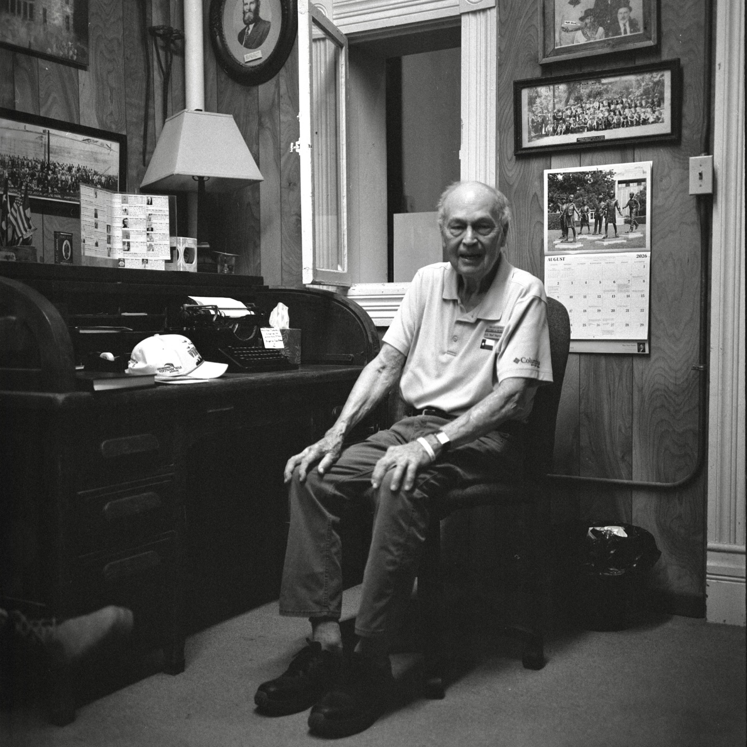 A black-and-white film photo. Bud, a volunteer, is seated at a desk in the old Milam County Jail in Cameron, Texas. Saturday, August 29, 2026.