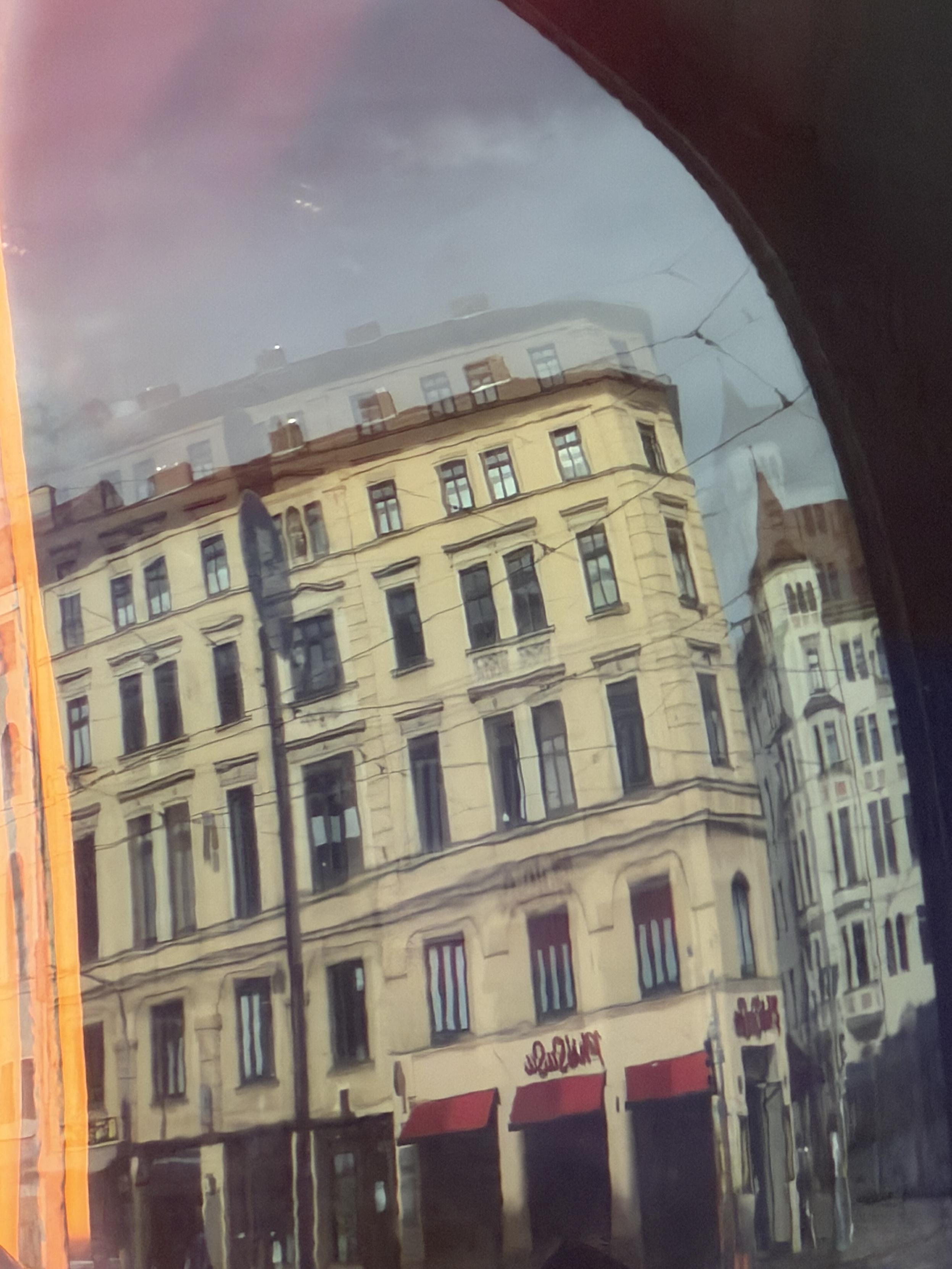 Street scene photographed through a curved, slightly cracked pane of glass, showing a pale yellow historic corner building with multiple rows of windows and red awnings at street level. Graffiti lettering is visible along the lower part of the building. In the background on the right, another white building with a turret. The top of the frame shows an overcast sky, with an orange light reflection running along the side edges, and a curved dark area darkening the upper right corner.