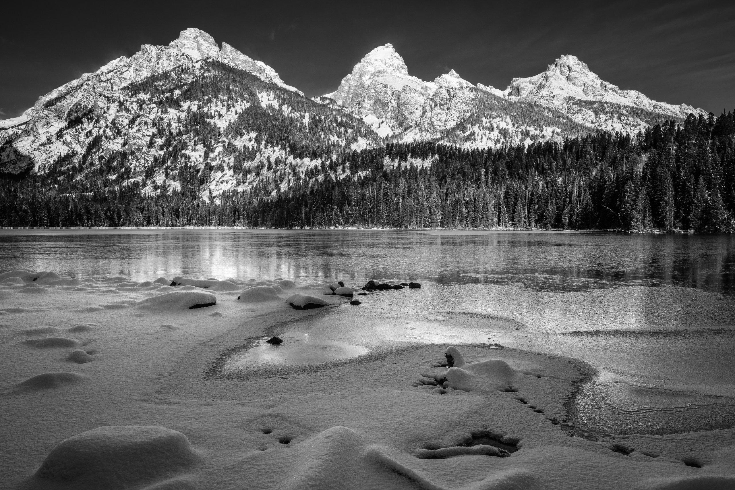 Logs and rocks covered in snow on the shore of a frozen Taggart Lake. In the background, Nez Perce Peak, Grand Teton, Mount Owen, and Teewinot Mountain.