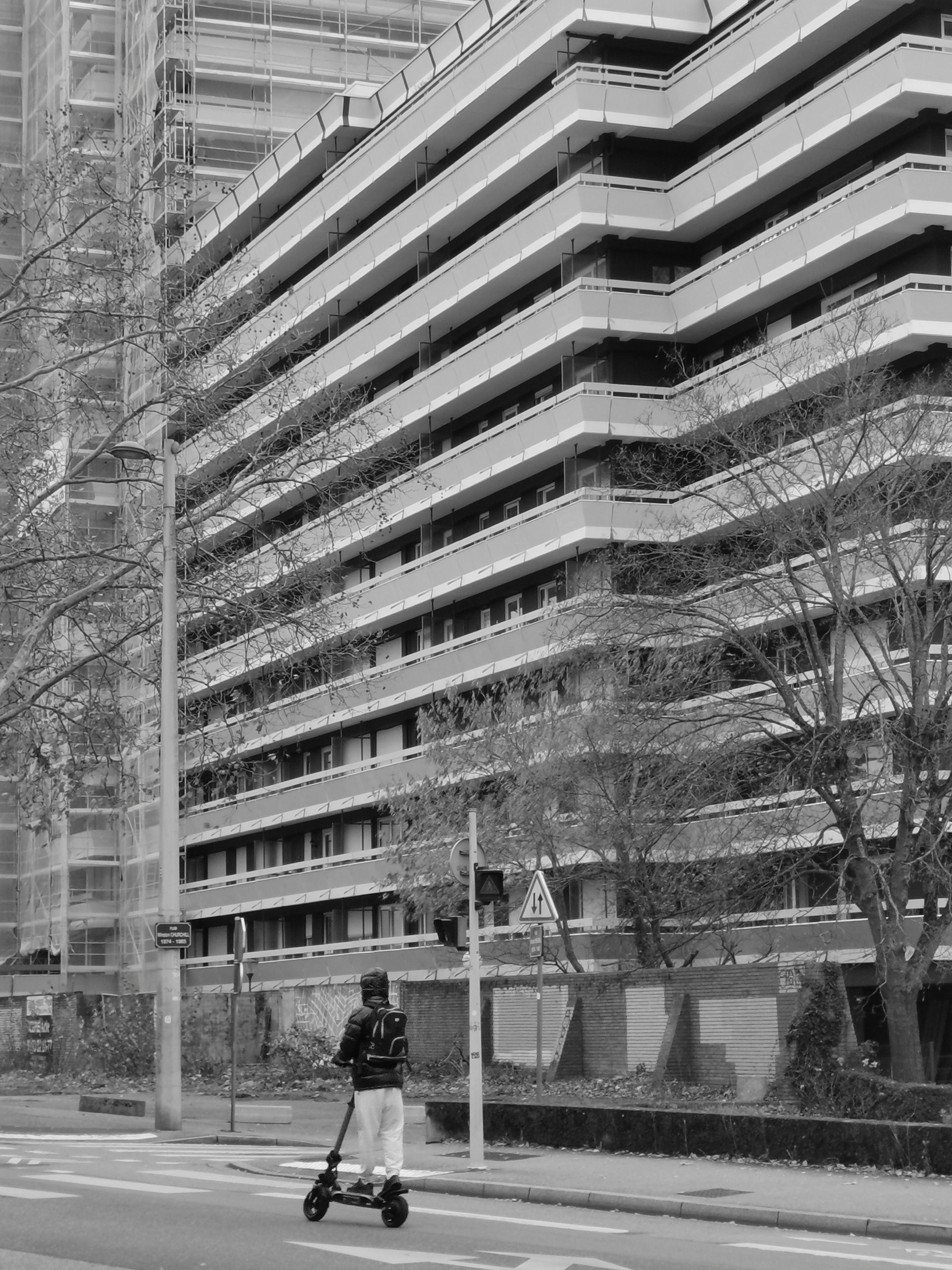 Une personne en trotinette passe devant la vaste façade d'un immeuble massif.
