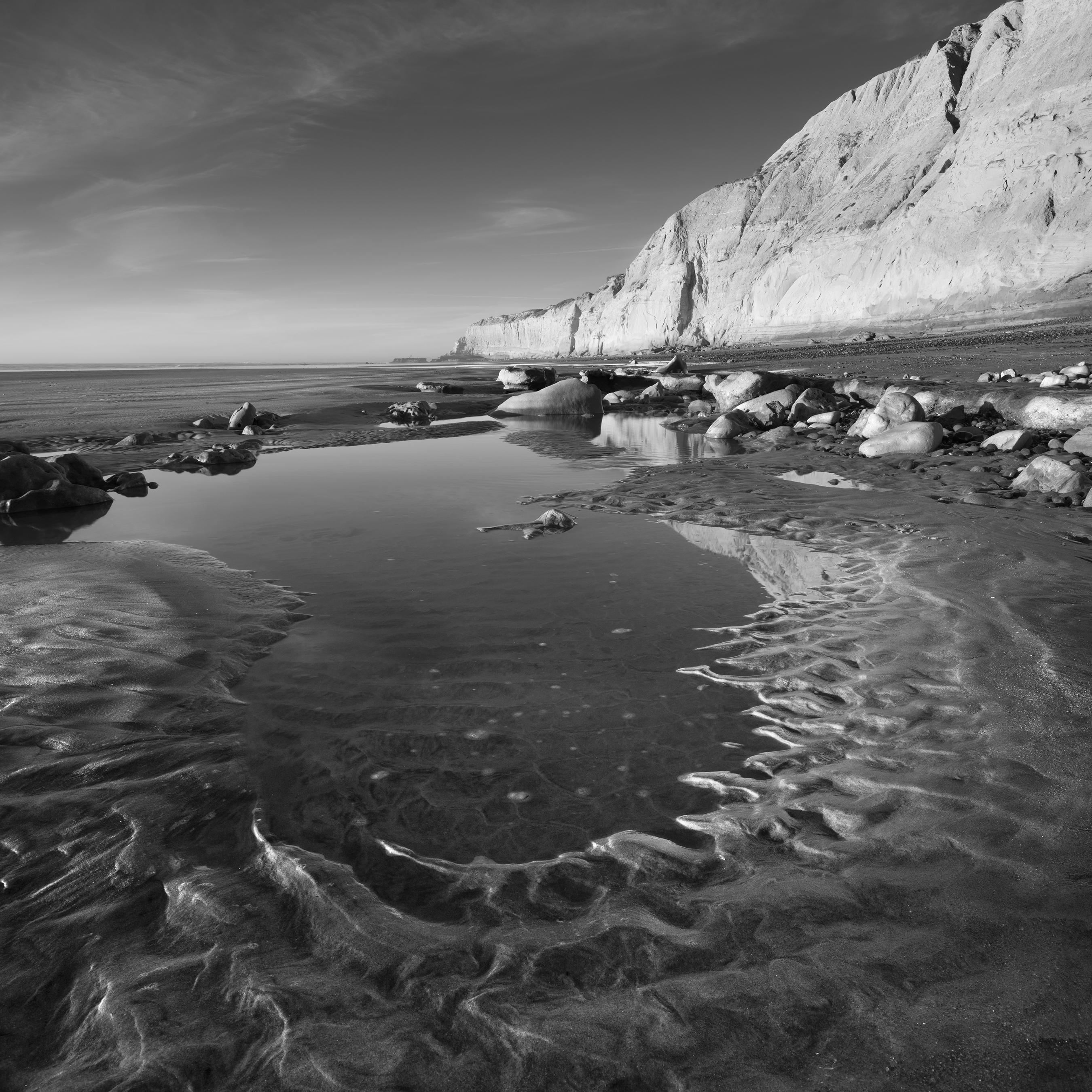 A black and white photograph of a pool of water on the beach at Torrey Pines, San Diego, California.  Beach cliffs rise tall in the distance.