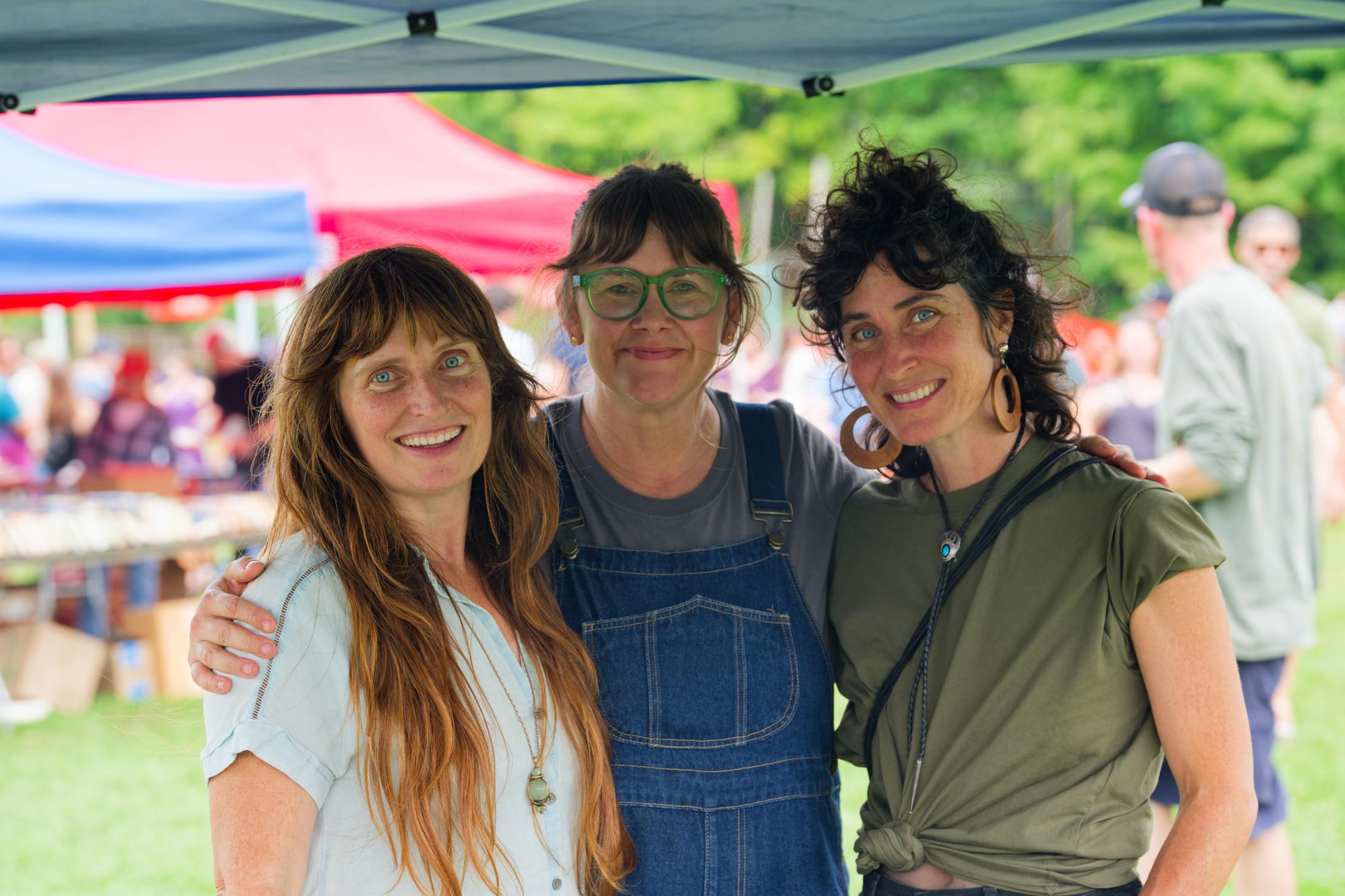 Three women from left to right with long wavy blond hair, short hair and suspenders and wavy dark hair with a brown shirt with a much brighter environment of blurred out people and tents and stuff behind