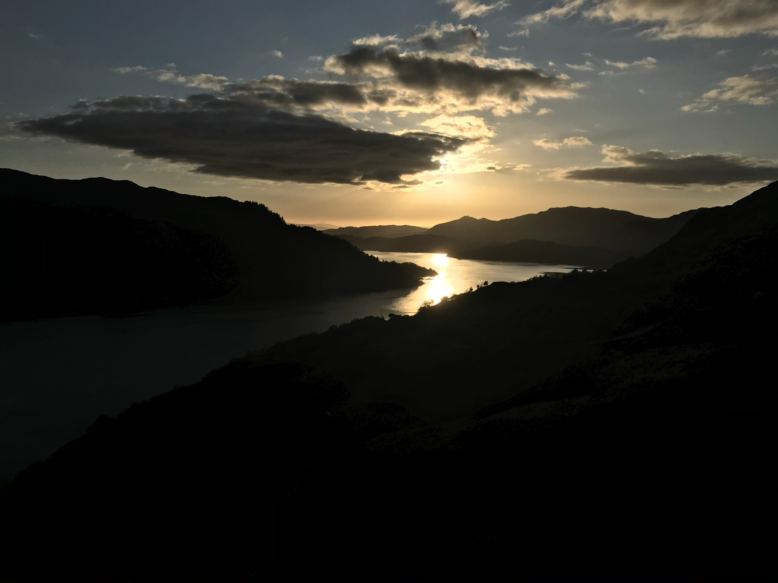 The sun setting over a Highland landscape in the north west of Scotland. The sky a mixture of yellow and blue, cushioned by clouds over the dark, moody, brooding hills.