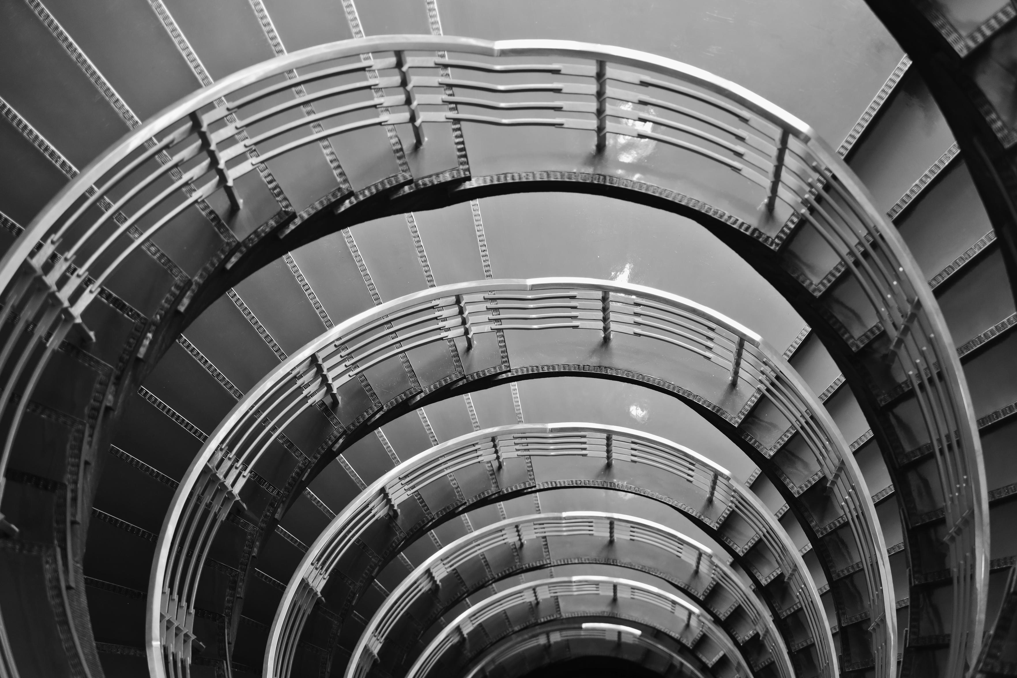 Schwarzweiss-Foto eines Treppenhauses von oben nach untengesehen. Die Ausführung des Handlaufs sowie der Treppenstufen kann als Industrial-Look beschrieben werden. Das Treppenhaus ist menschenleer.