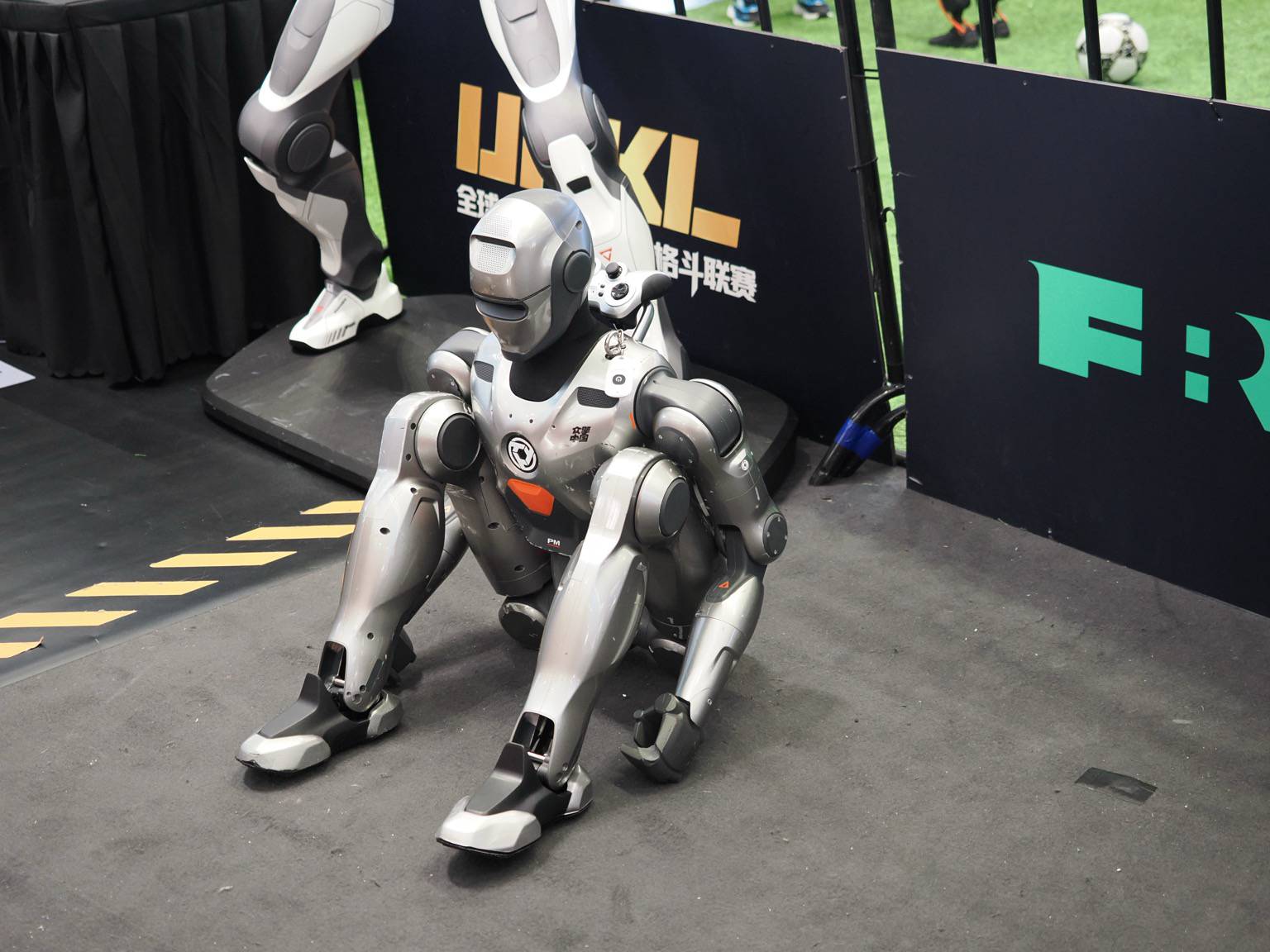 A robot sits on the ground with its knees pulled up to its chest. At the back of its head is a game controller and behind it are the legs or another robot