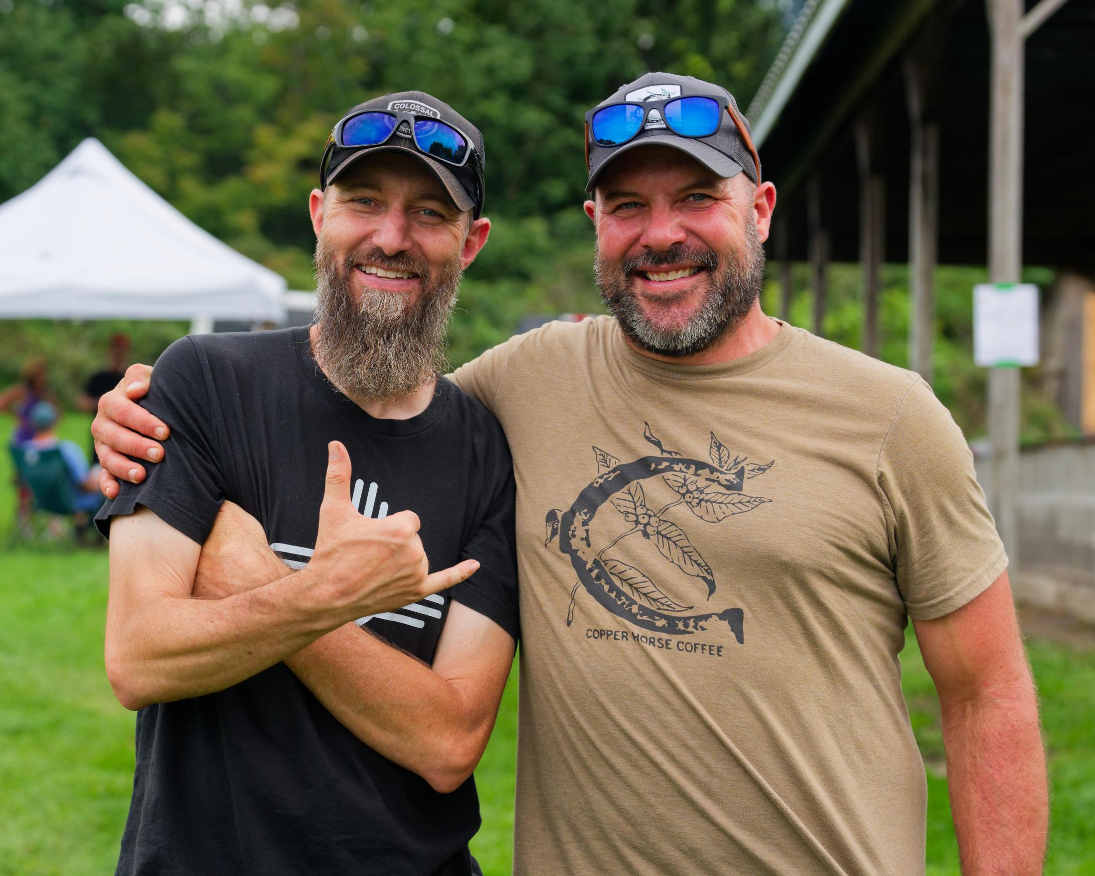 The guy on the left has a black shirt with a new mexico logo and is making a hand sign and the guy on the right has a light bround horseshoe t-shirt,  both have smiles, beards, baseball caps and glasses on top of their caps;  behind them a hedgerow, a tent, and a chicken-cooking structure
