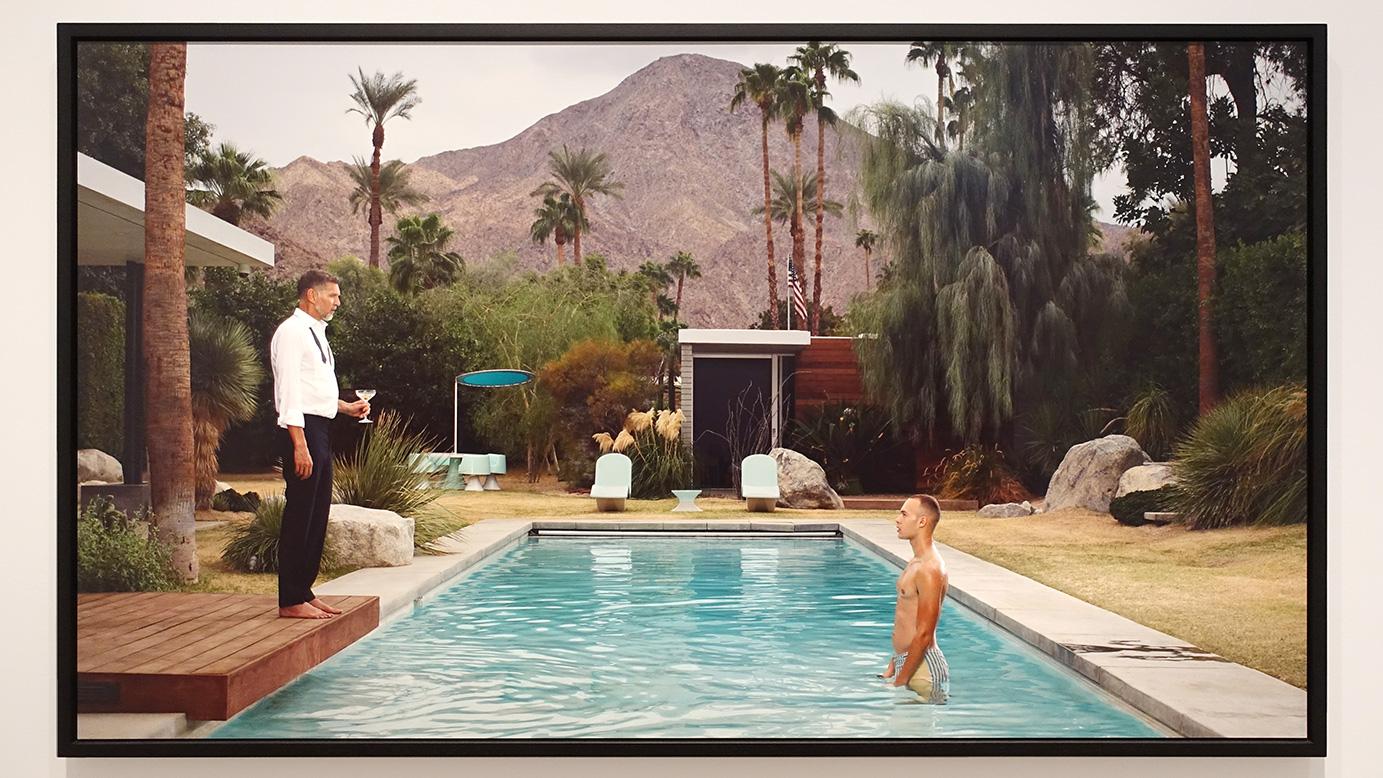 Een kleurenfoto uit de serie Palm Springs uit 2018. Een foto van een privézwembad omringd door tropische planten en bomen en op de achtergrond een hele hoge kale berg. In het zwembad staat het model Alex rechtop tot aan zijn kruis in het water. Op de houten vlonder staat Erwin Olaf zelf. Zwarte broek, wit hemd en een losgemaakt zwart strikje en blote voeten alsof hij net terug is gekomen van een sjieke samenkomst.
In zijn hand houd hij nog een champagne-glas vast. Hij kijkt het model in strak aan. Die kijkt hem ook strak aan. De sfeer is een beetje surreëel. Is er een spanning tussen de beide heren? Is het liefde?