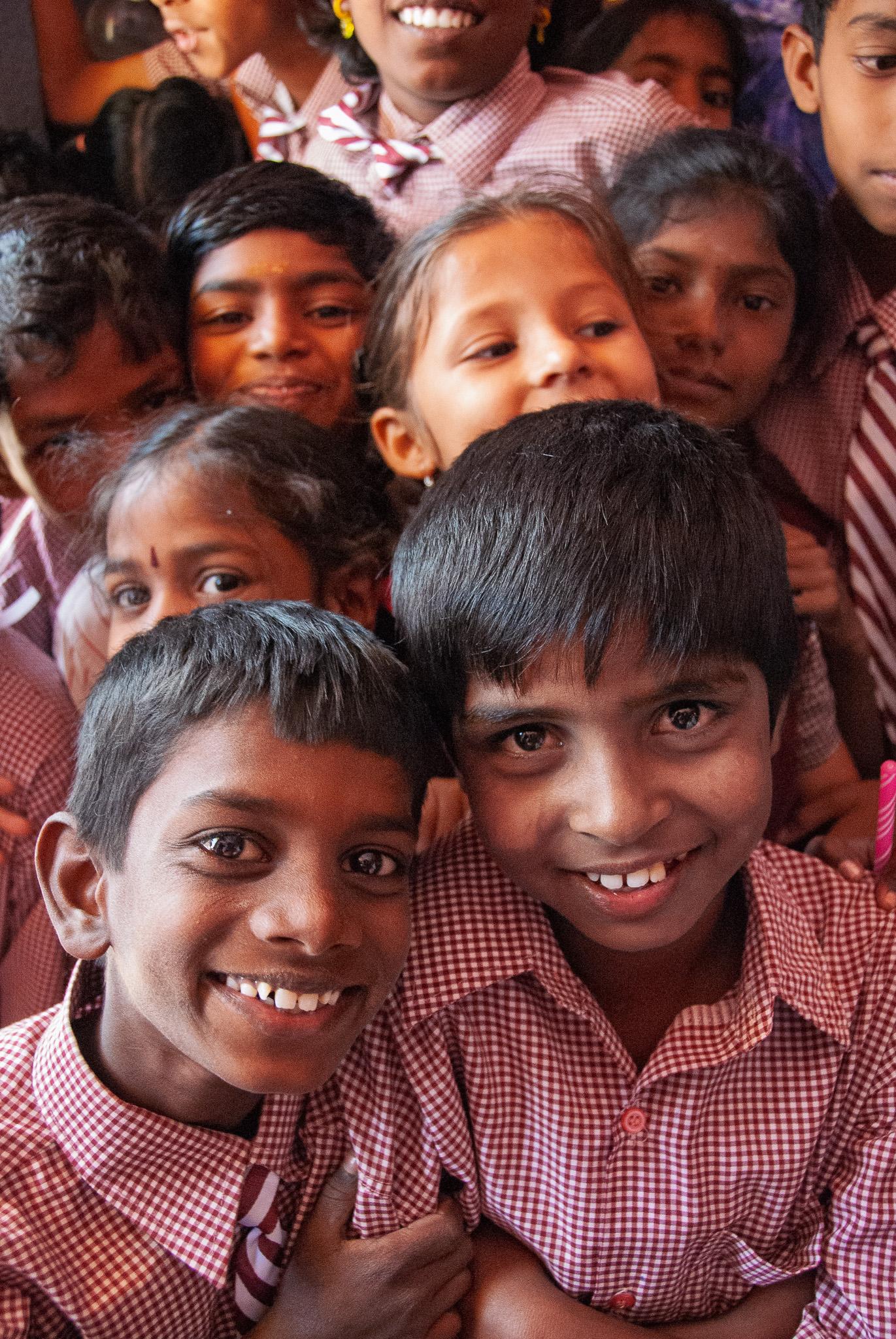 A close crowd of smiling schoolchildren in red checkered uniforms fills the frame, with two boys in front and several girls and boys behind them.