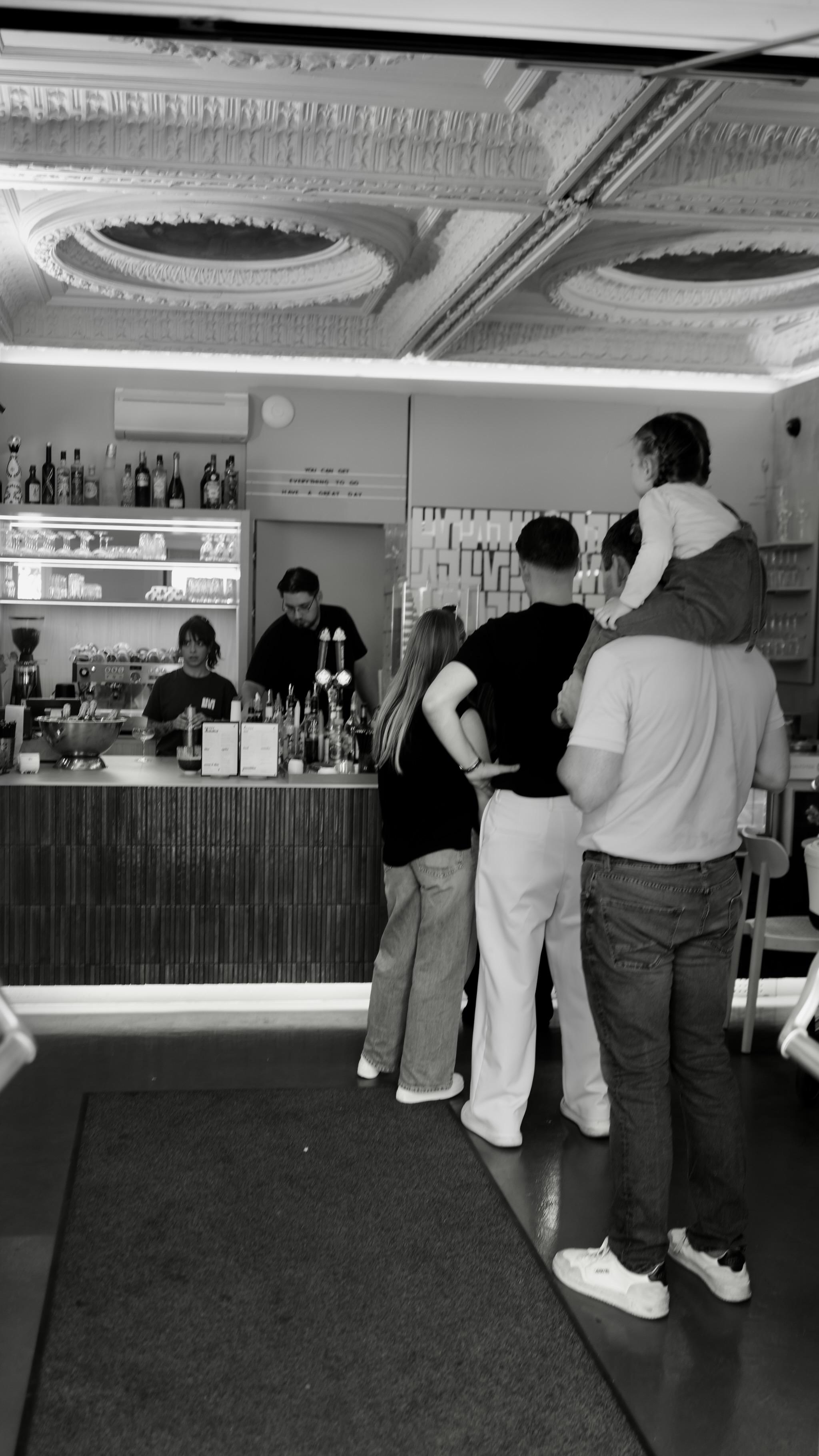 Picture is taken in a coffee bar. Two people are behind the counter, working there and looking to the customers.
In front of them is a line of people. In the first row is a girl, looking to her right as she seems to speak to someone. Behind her is a guy, tall with black shirt and white trousers. Last In line is a man with a kid on his shoulders. 
Photo is black and white.
