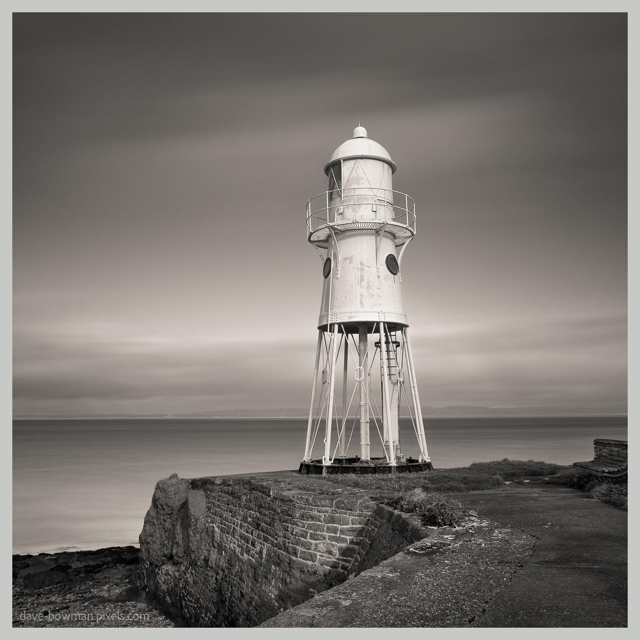 A photograph of Black Nore Lighthouse standing above the rocky shoreline at Portishead in Somerset, England. The distinctive white lighthouse, built in 1894, rises prominently against a softly blurred sky, while a long exposure has smoothed the water around the rocks. The monochrome treatment gives the scene a calm, timeless appearance.