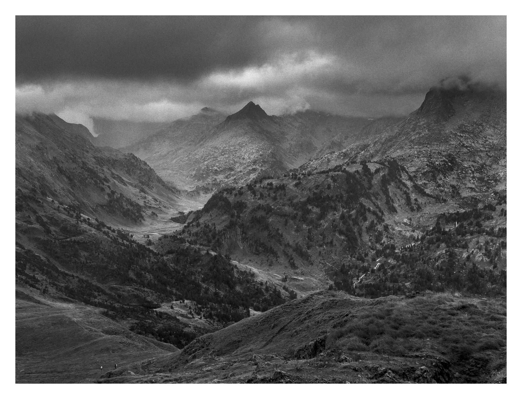 Mountain landscape under a dark overcast sky. In the foreground, a dark hillside covered in grass slopes towards the bottom left corner, with two tiny human figures on a trail on the bottom left, providing a sense of scale against the vast landscape. In the mid-section, rugged ridges covered with scattered trees and rock formations, with a deep valley winding through it. The overall tone is dark, but light filters unevenly across the clouds, creating pockets of light in the landscape. The background features a prominent triangular peak, flanked by steep, jagged mountain walls on both sides. The rest of the peaks and ridges appear cloaked under low, heavy storm clouds.