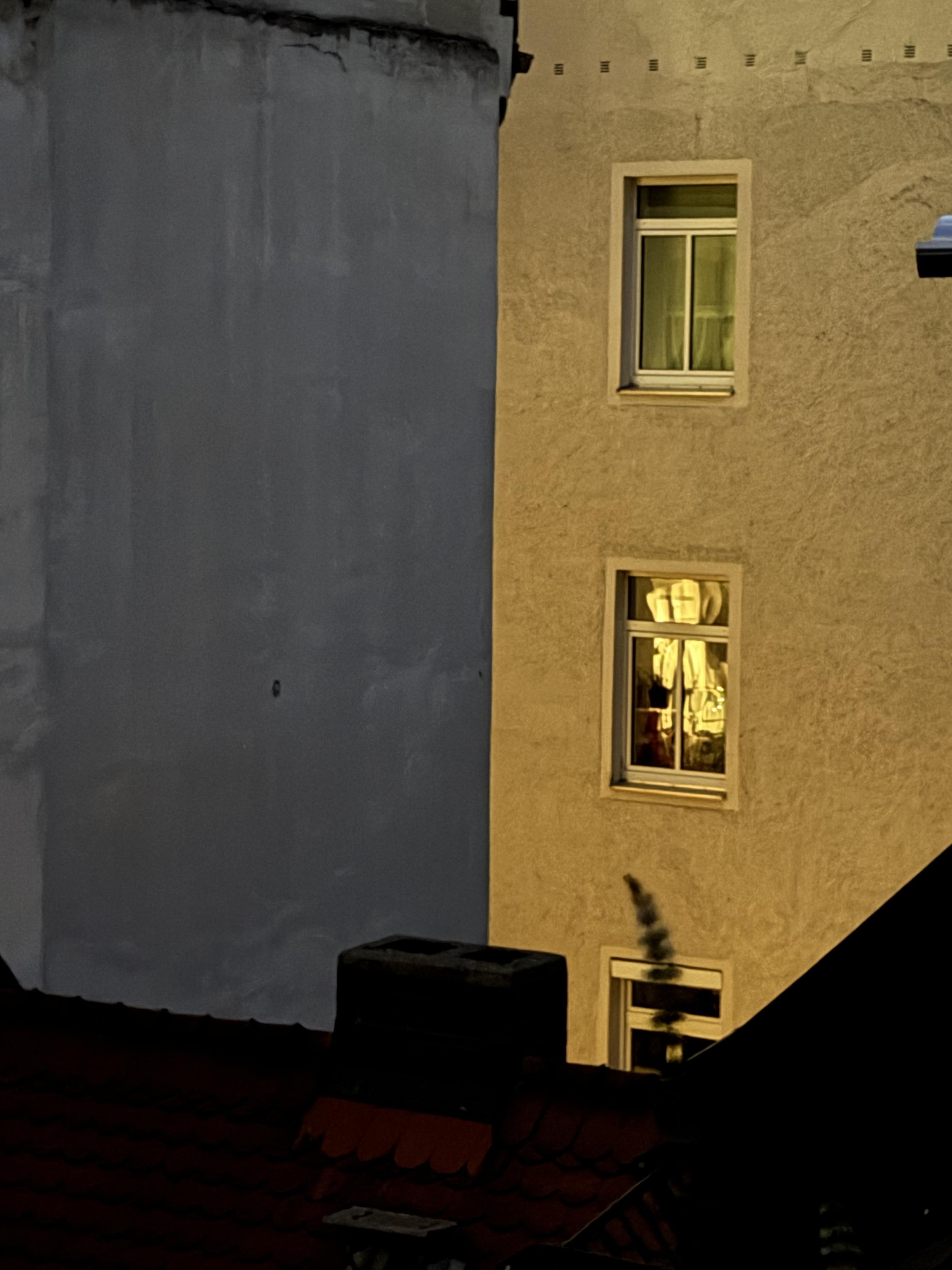 Nighttime shot of two adjoining building façades. On the left, a smooth grey wall; on the right, a warmly lit golden façade with two white-framed windows stacked above each other — the upper one shows a bright curtain, the lower one reveals objects and draped fabric. Below them, a third, darker window with a blurred dark shape in front of it. At the bottom, a red tile roof with a rectangular chimney lies in shadow.