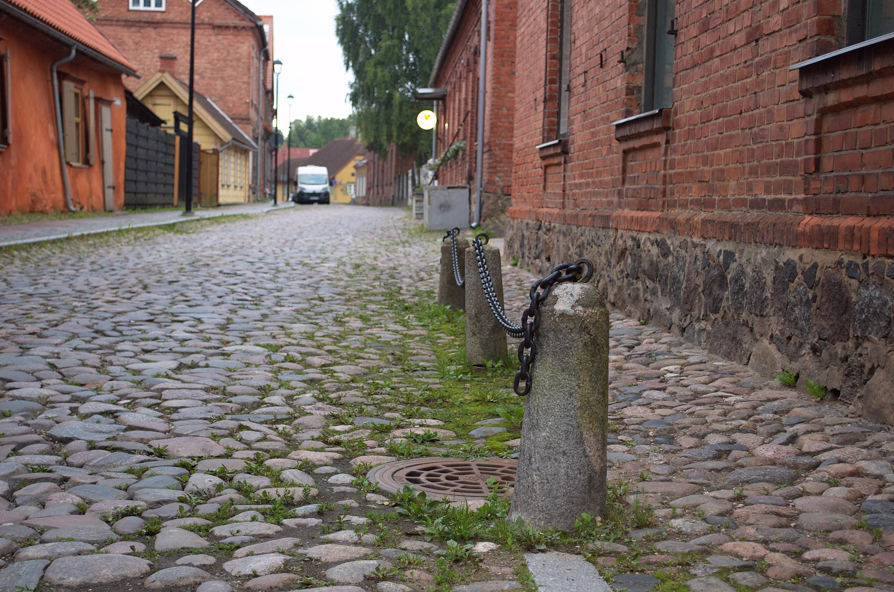 Photo looking along a narrow street paved with irregular round cobbles. The houses visible are more than a century old, built from red brick or wood. One low house covered in bright orange plaster is visibly leaning away from the street. Along the left side of the street runs a sidewalk paved with granite slabs. On the right are three granite bollards, connected together with black-painted iron chains. Around the bollards, grass is growing between the cobbles.