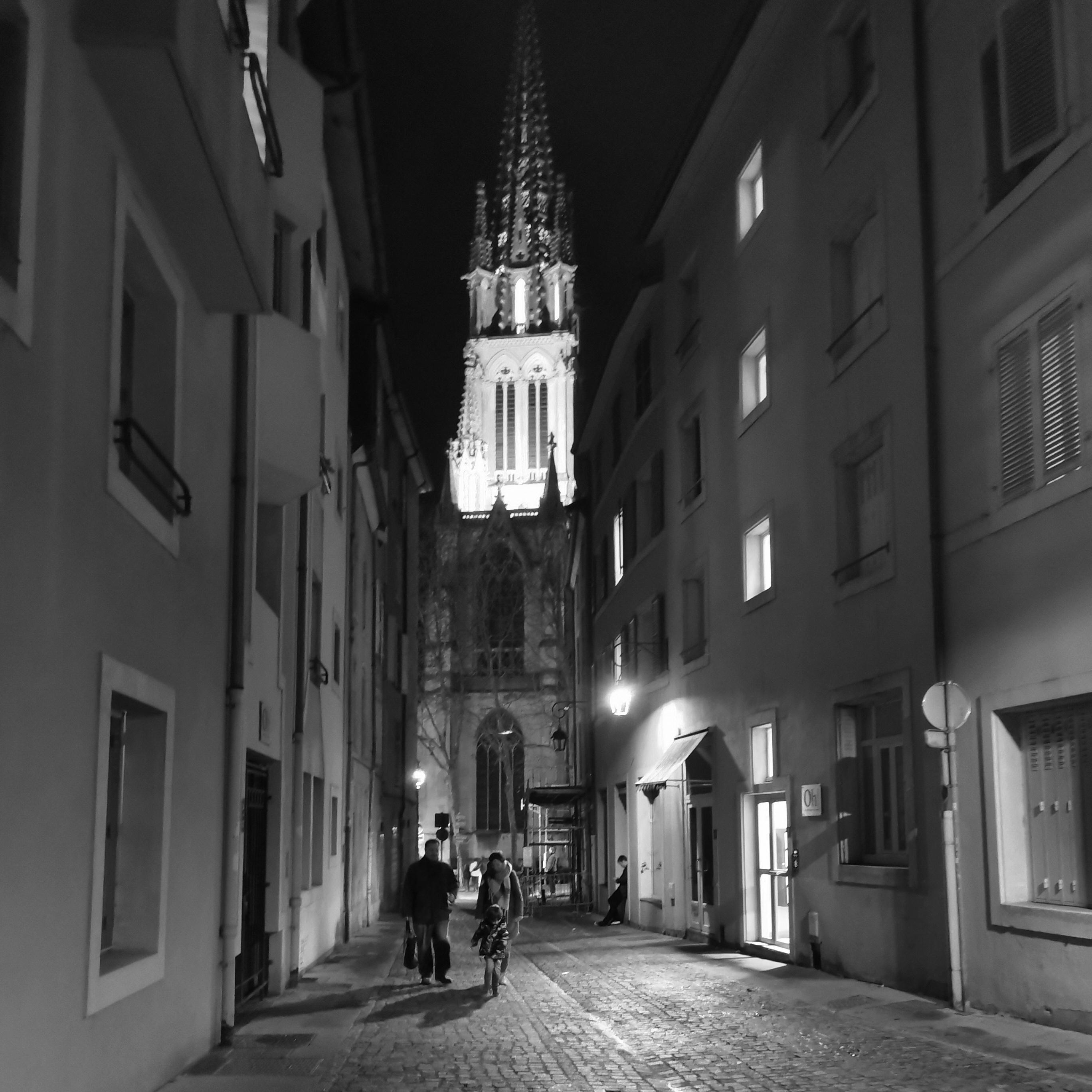 Dans une rue de la vieille de Nancy, un groupe de personnes, en fond le clocher éclairé de l'église Saint-Epvre.